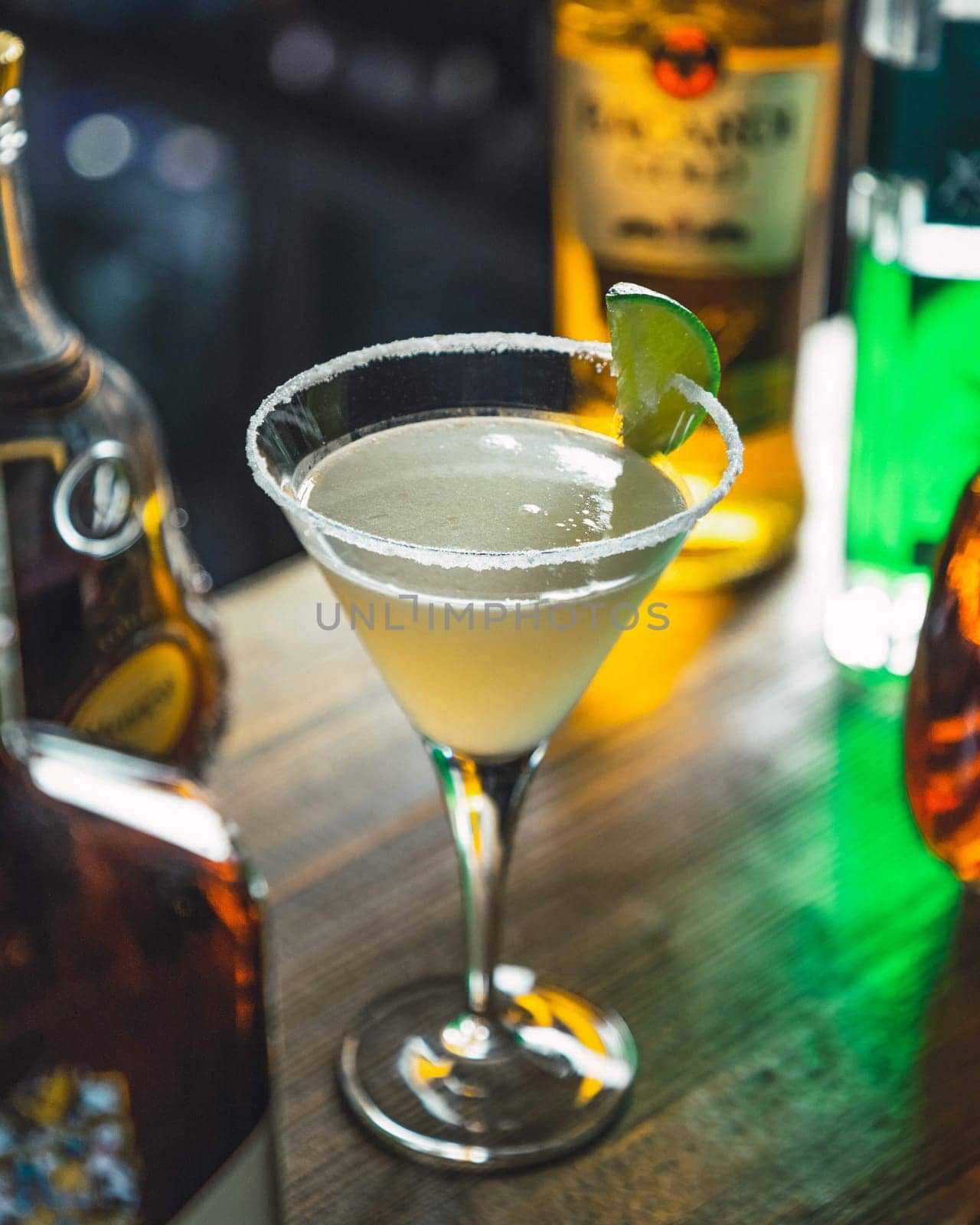 Cocktail glass with a salted rim, a translucent yellow drink, and a lime wedge is standing on a bar counter