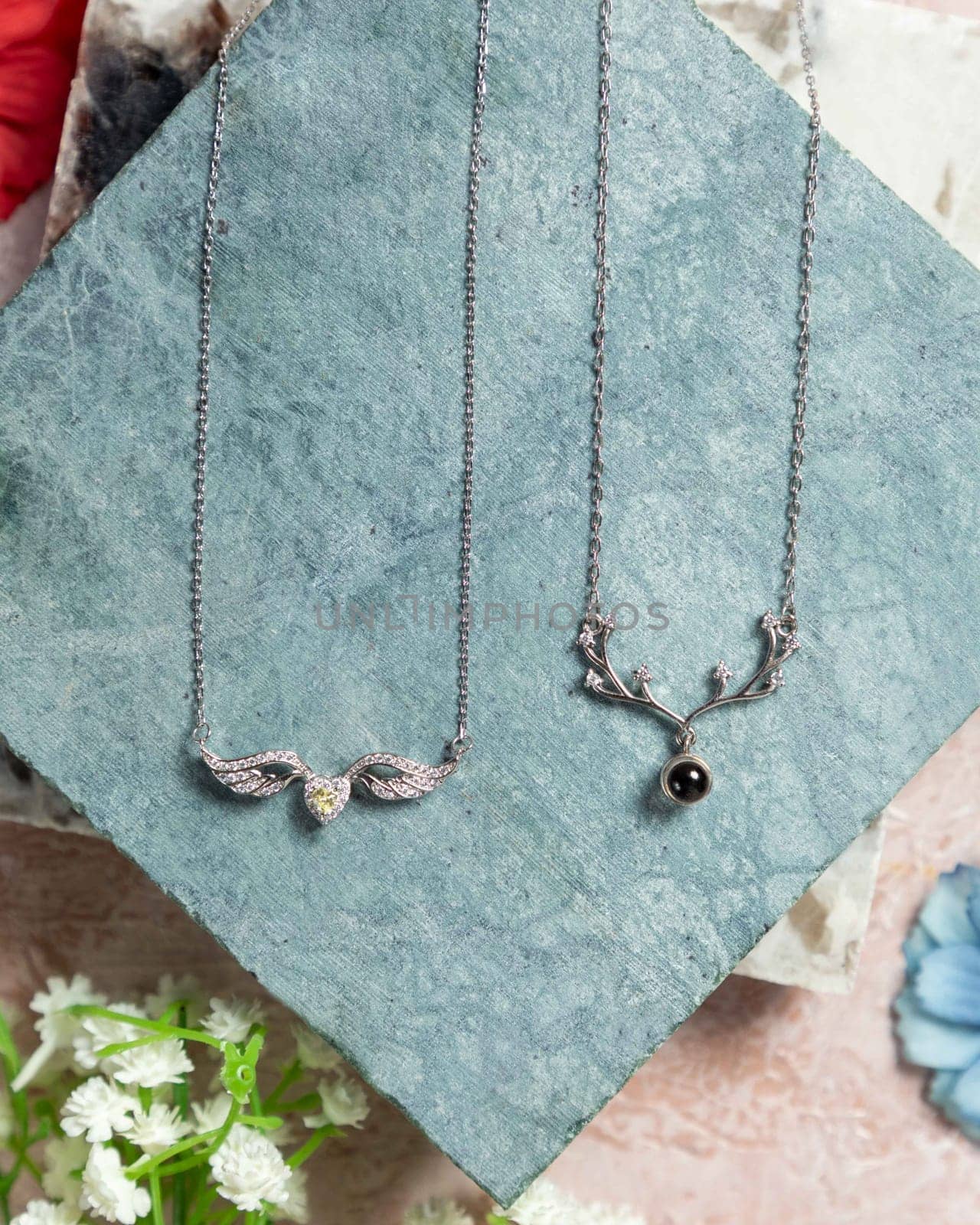 Two silver necklaces on textured stone, surrounded by flowers, showcasing their elegant design