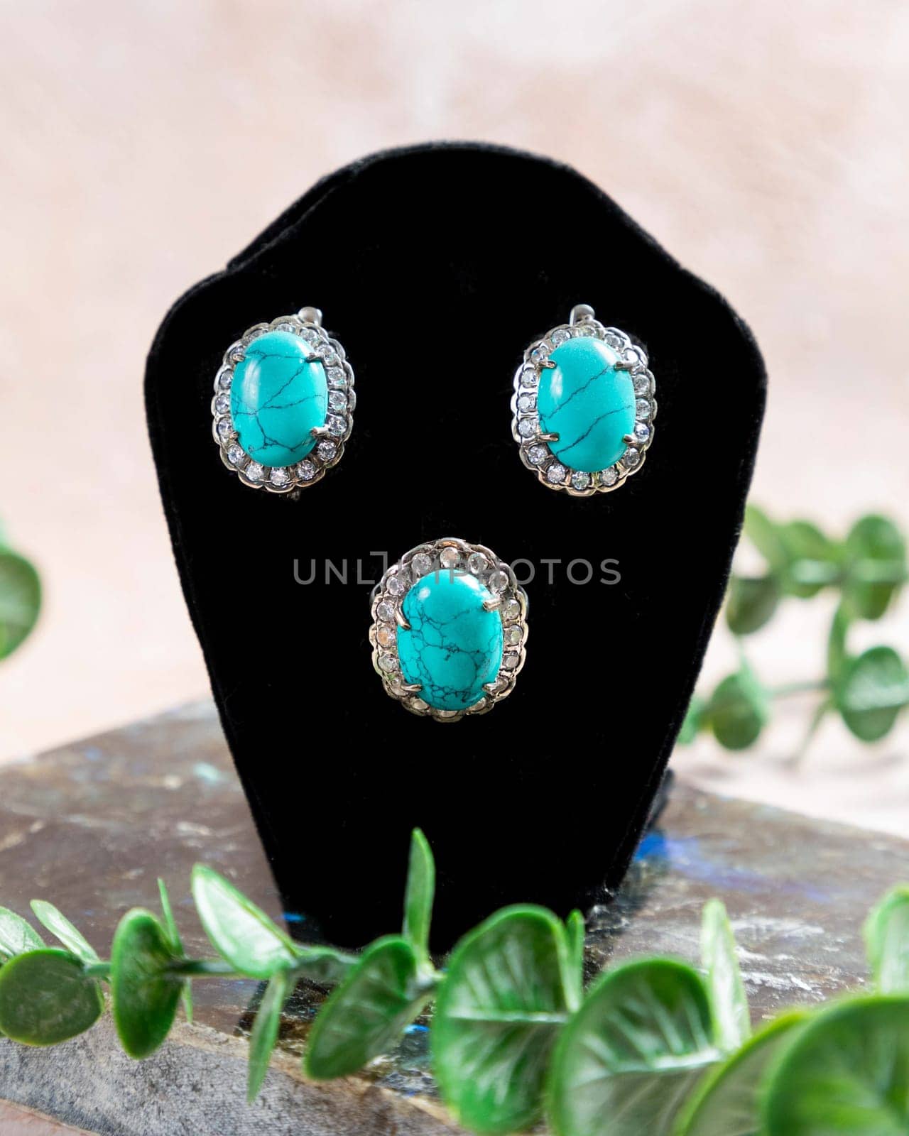 Silver jewelry set with a turquoise stone and sparkling diamonds is displayed on a black stand with green plants in the background