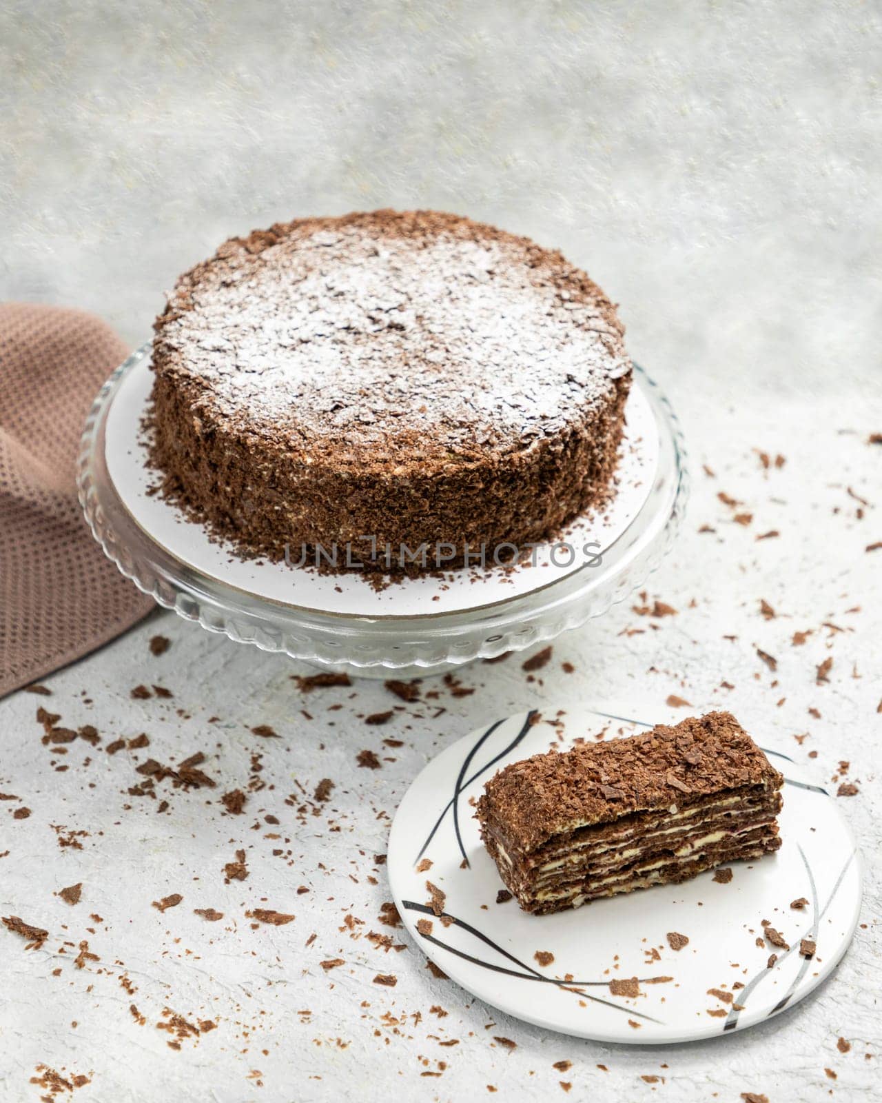 Chocolate cake with chocolate chips sprinkled with powdered sugar standing on a glass stand with a slice on a plate