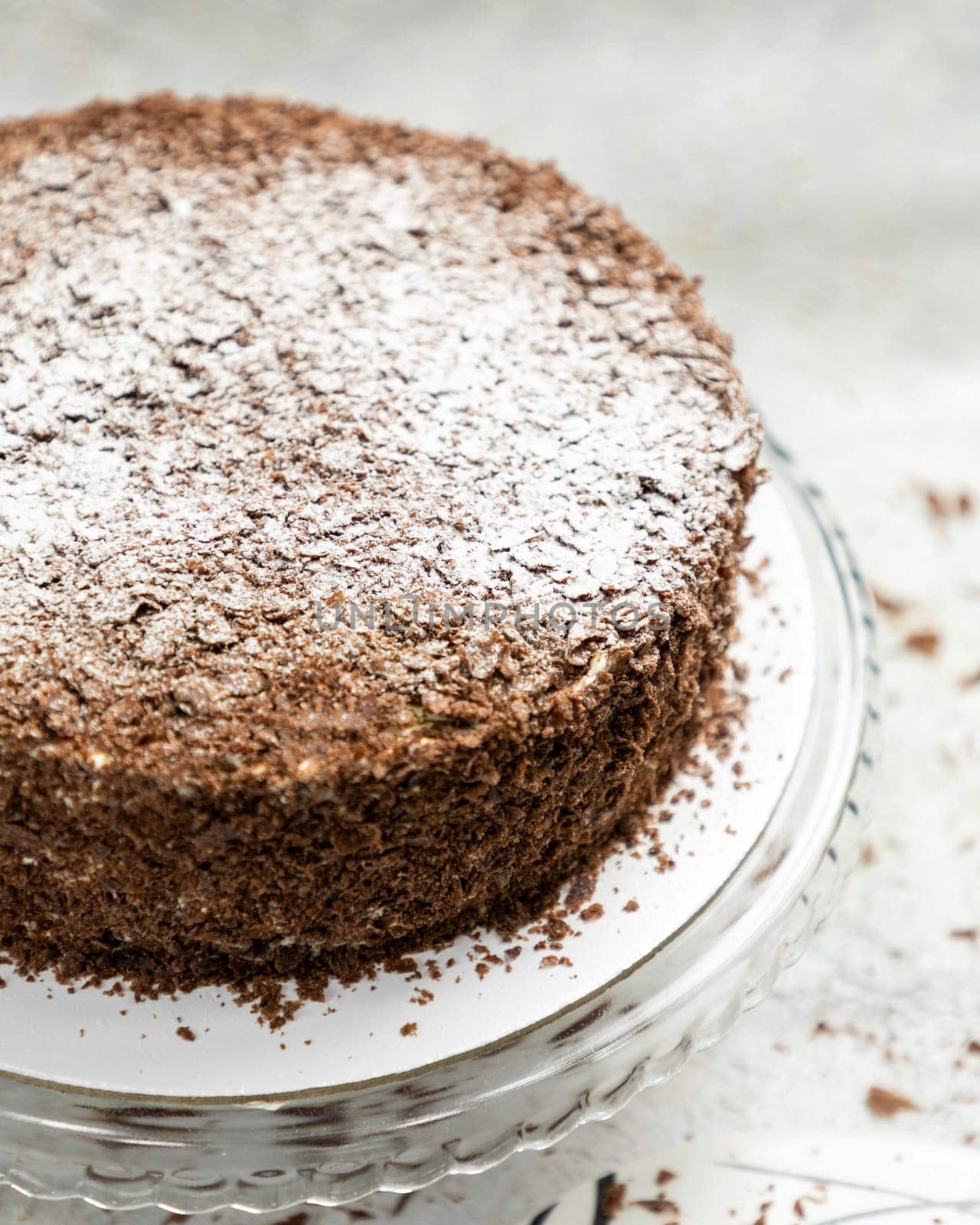 Chocolate cake with chocolate shavings and powdered sugar is sitting on a glass cake stand