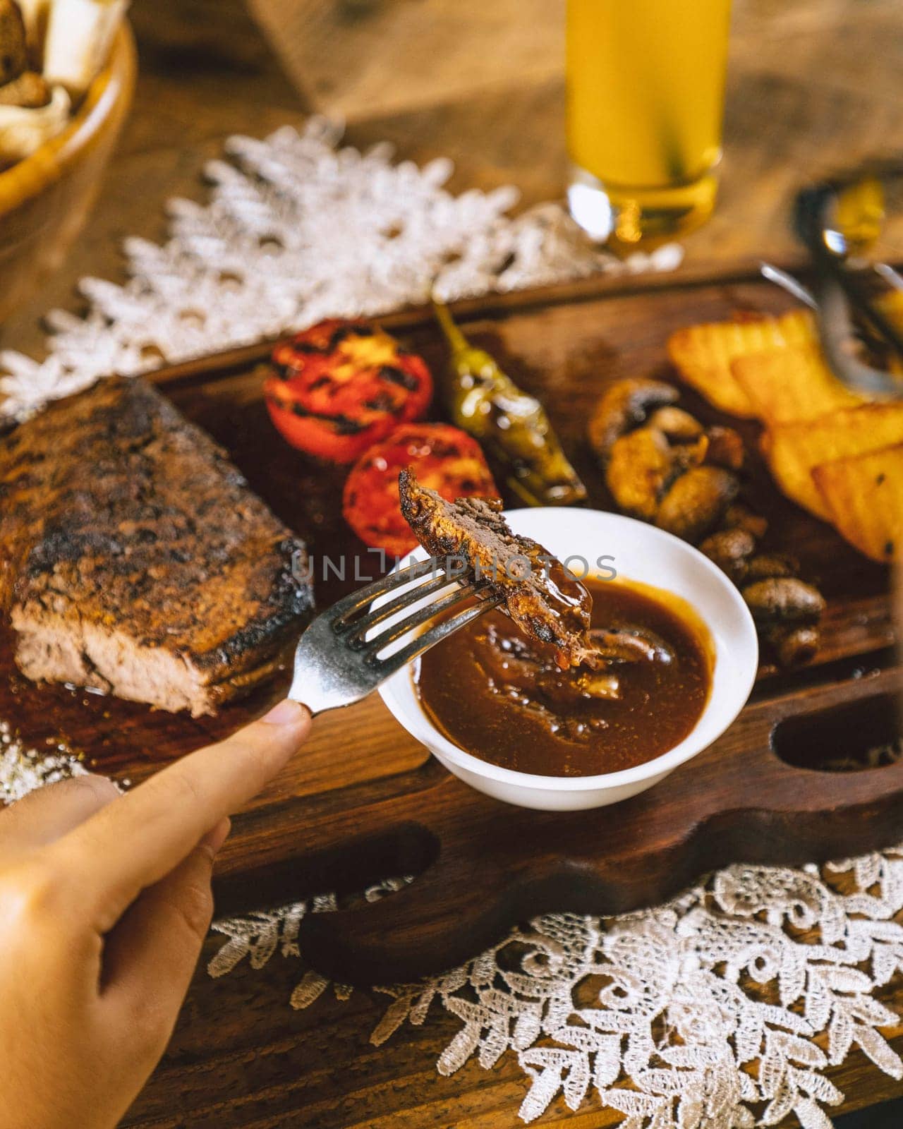 Person is dipping a piece of grilled steak into a bowl of sauce, with a glass of beer in the background