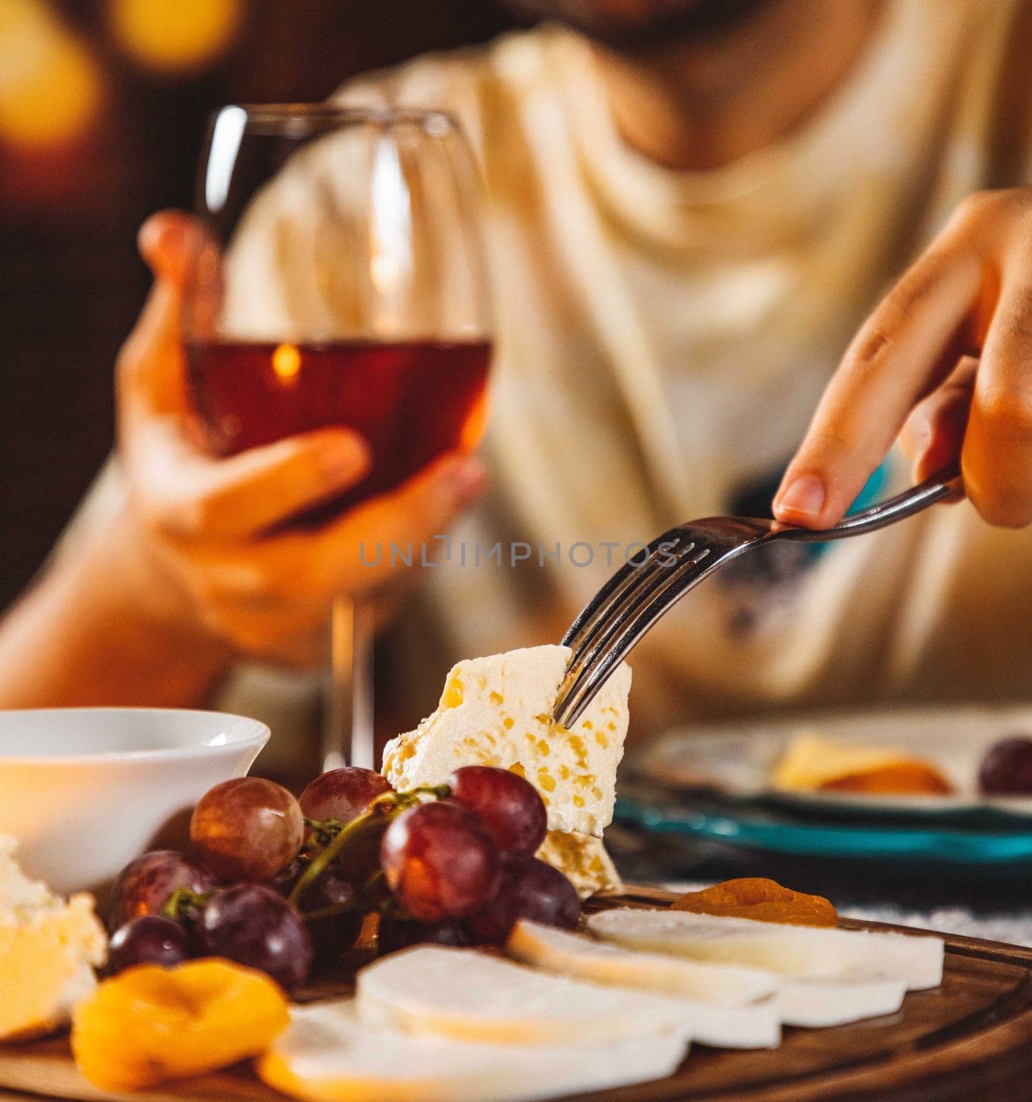 Man picking up a piece of cheese with a fork from a cheese board with grapes and dried apricots, holding a glass of red wine