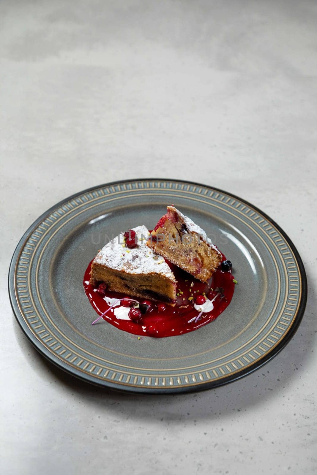 Two slices of delicious cake lying on grey plate with red fruit sauce and decorated with powdered sugar and red berries