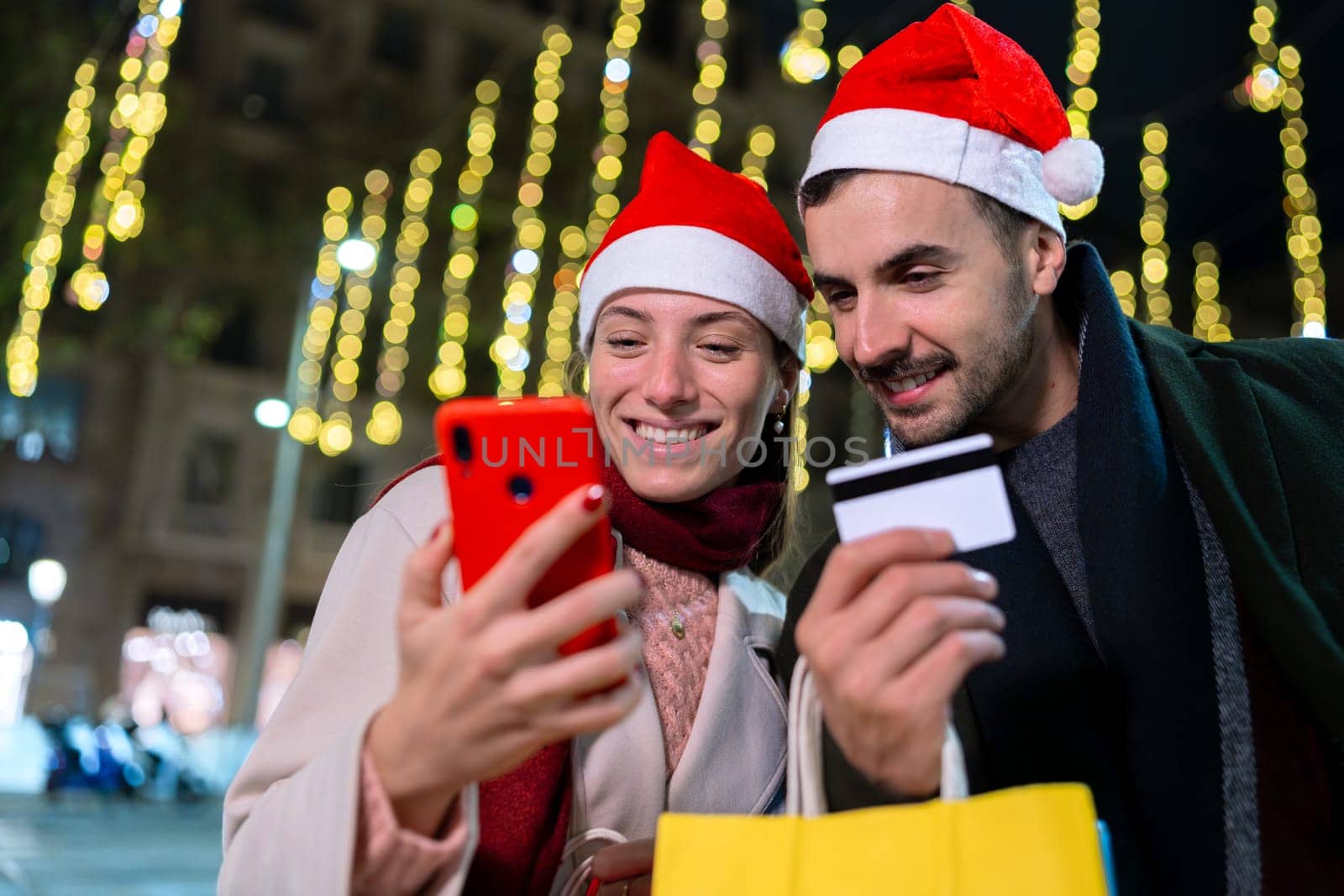 Smiling couple wearing Santa hats, standing under festive Christmas lights. They look at each other warmly, creating a joyful holiday atmosphere. One holds a phone, capturing memories.