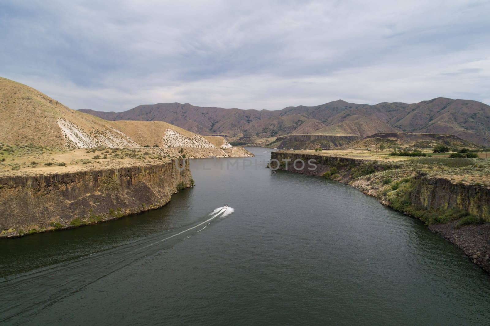 A boat is traveling down a river in a mountainous area. The water is calm and the sky is cloudy
