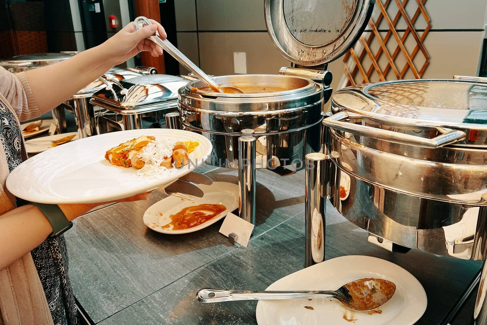 At the hotel's buffet table, a lady is scooping up food from a warming machine onto her plate.