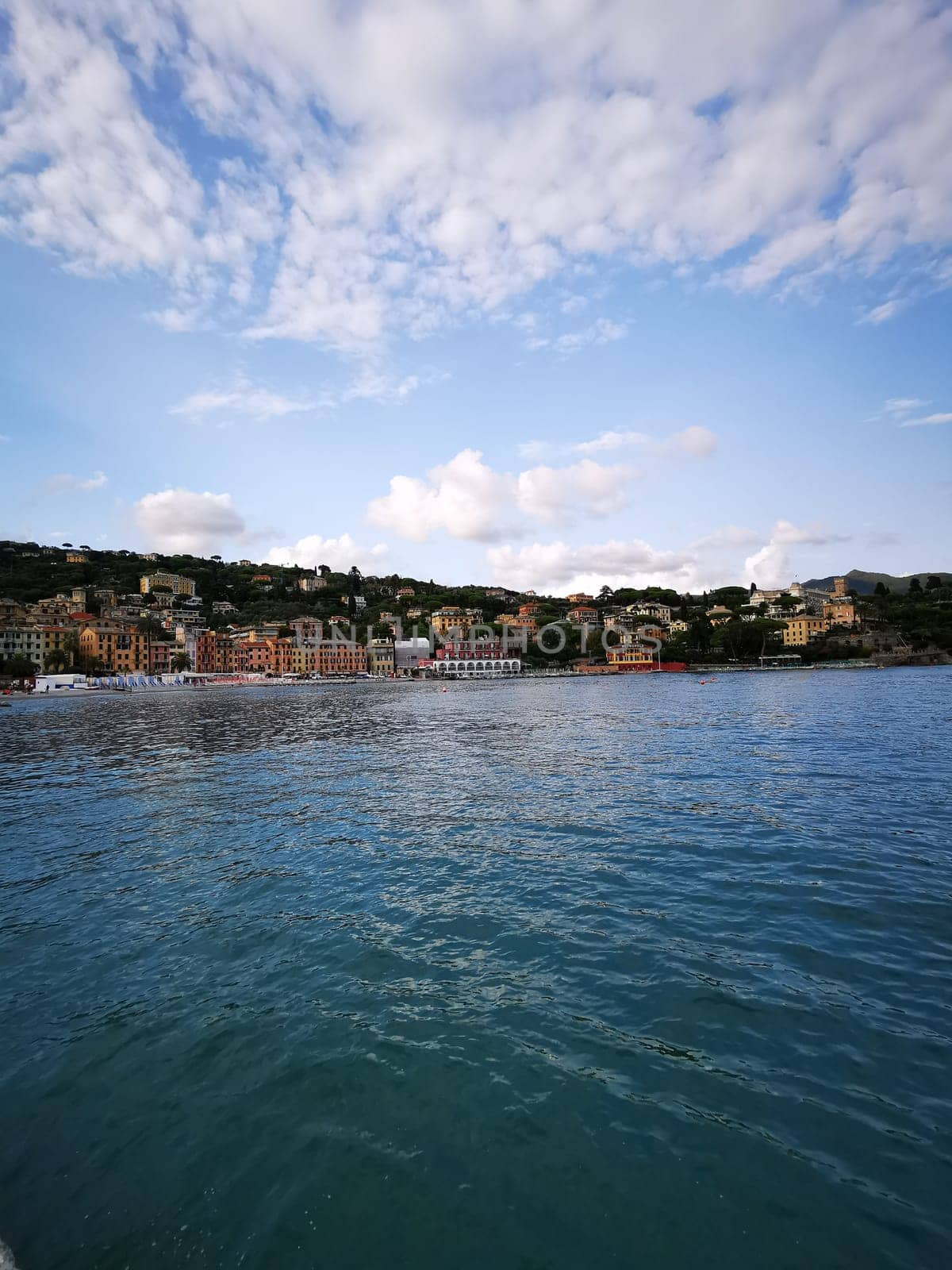 Colorful coastal buildings reflect off the calm water, surrounded by green hills and a bright blue sky, creating a serene seaside atmosphere.