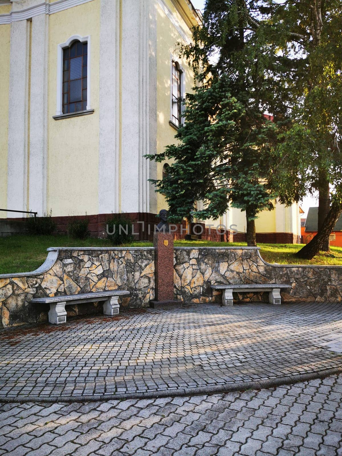 Stone benches provide a tranquil spot in front of a historic building, featuring a decorative bust surrounded by greenery on a bright day.