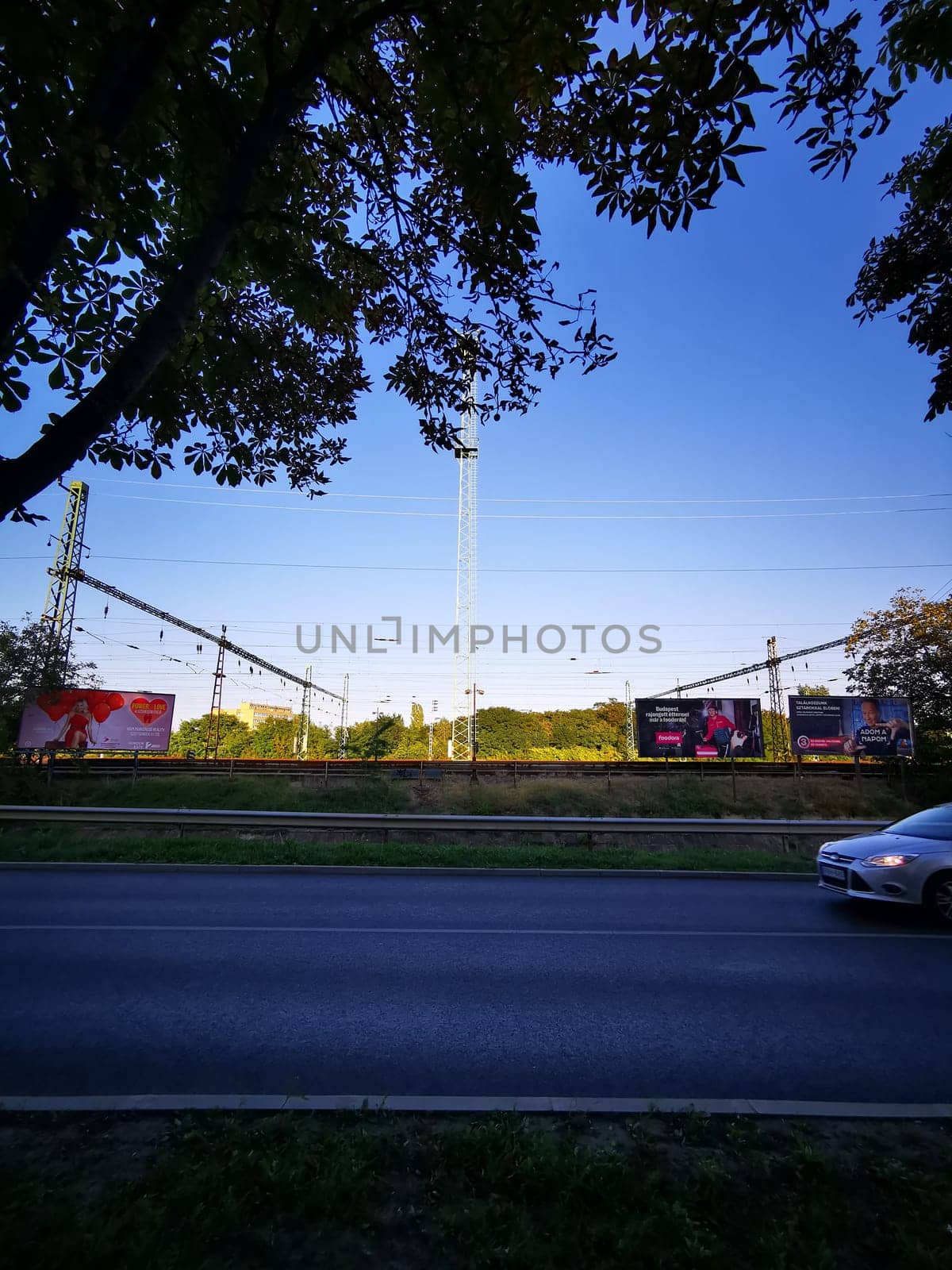 On a bright day, billboards line a busy road, showcasing advertisements while a car drives past the railway tracks, framed by lush greenery.