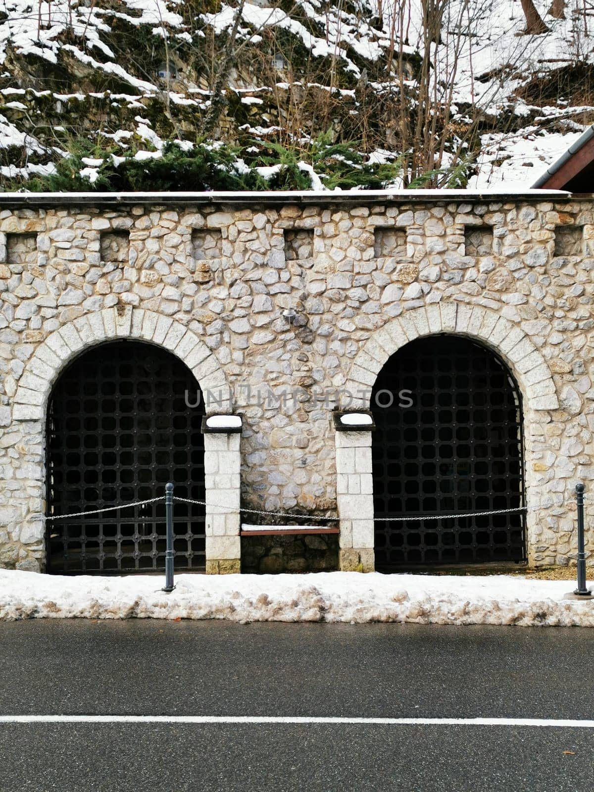 The solid stone structure features two arched entrances surrounded by snow, located beside a quiet roadway in a picturesque mountainous landscape.