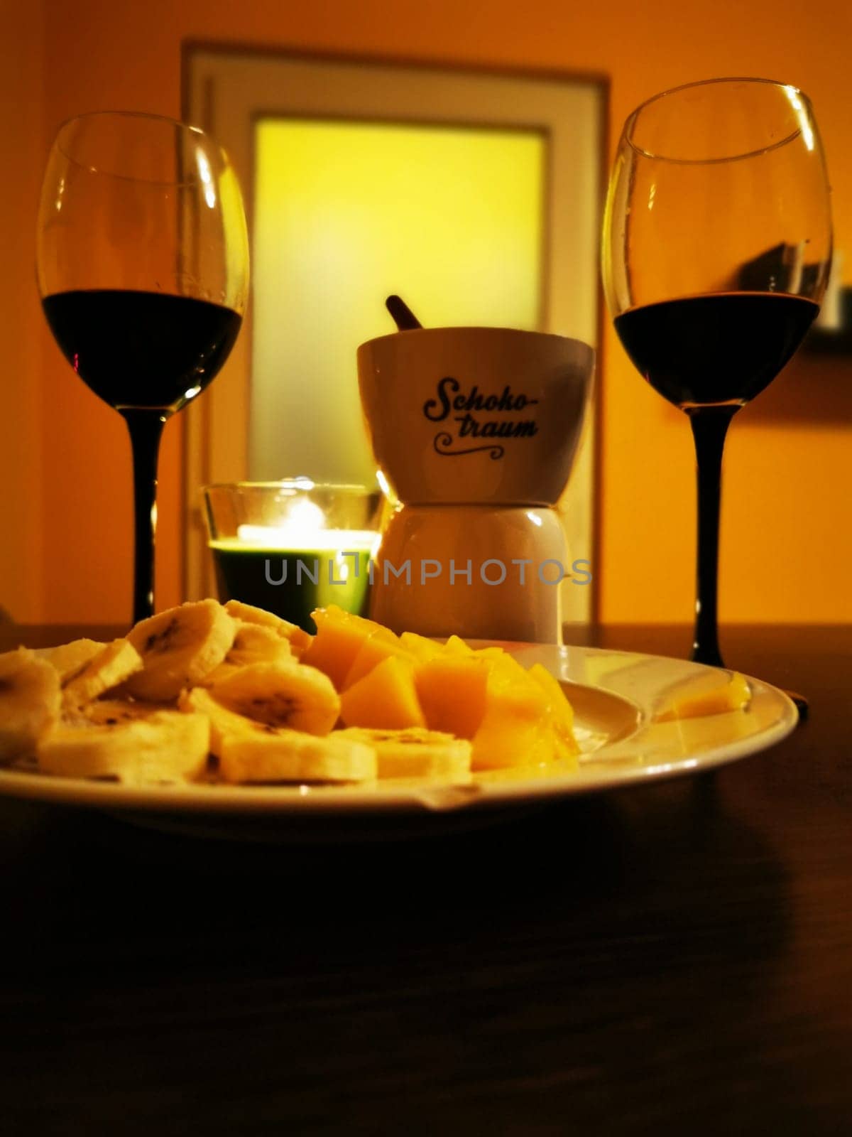 A dessert spread showcases chocolate fondue surrounded by sliced bananas and other fruits, paired with two glasses of red wine in a warm, intimate atmosphere.
