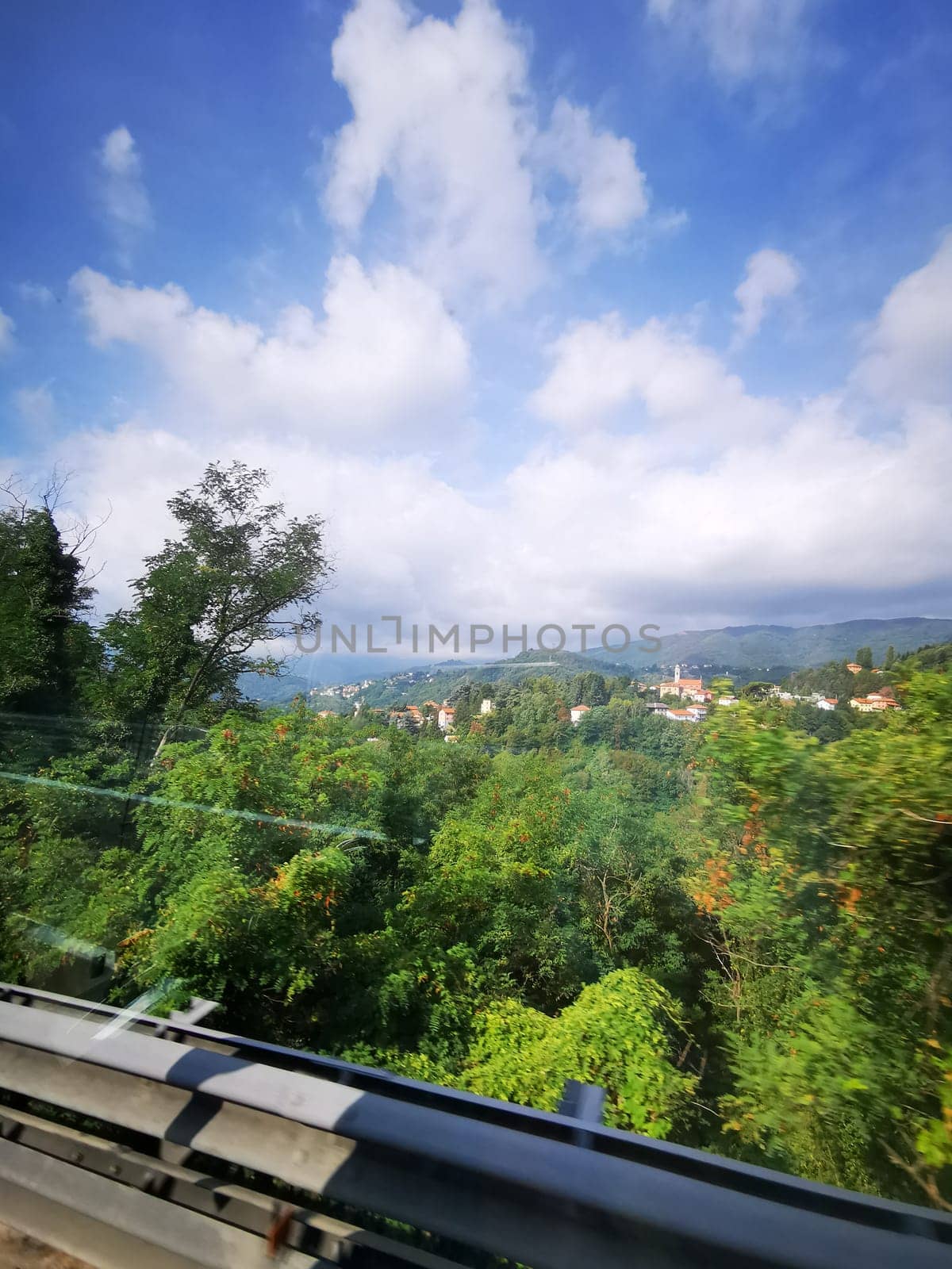 A vibrant landscape unfolds along a highway, showcasing green forests and distant hills beneath a blue sky filled with fluffy clouds.