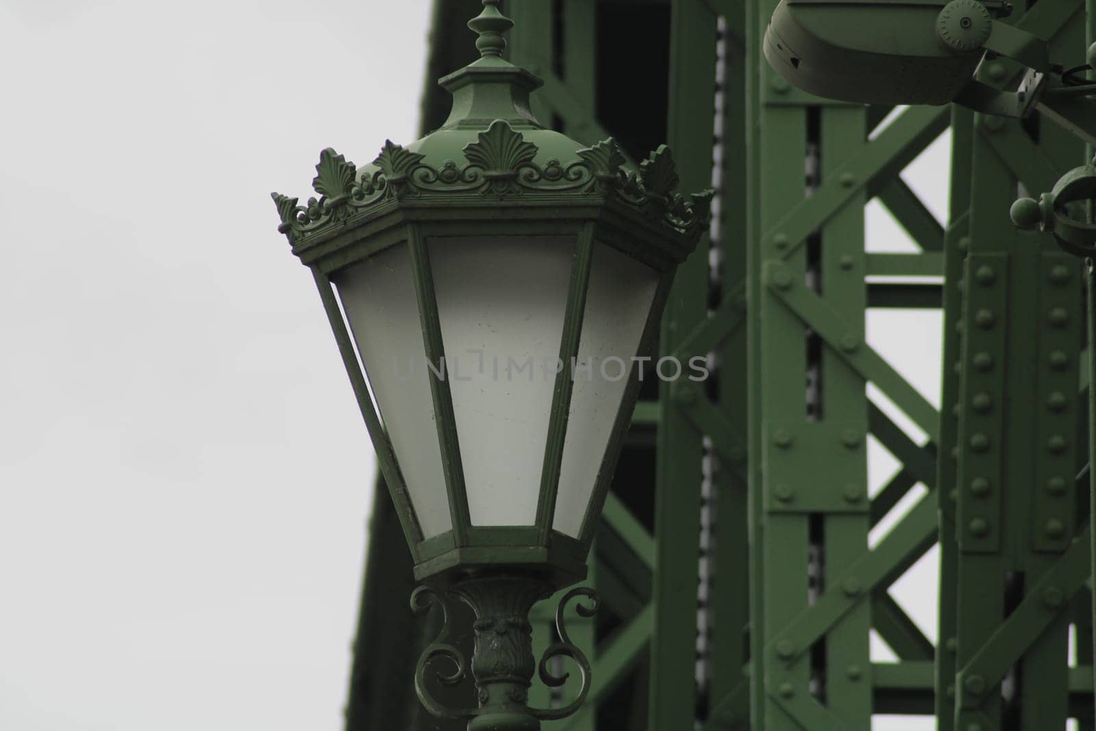An ornate green street lamp stands prominently, showcasing intricate designs while framed by a stark industrial structure, highlighting urban architecture.
