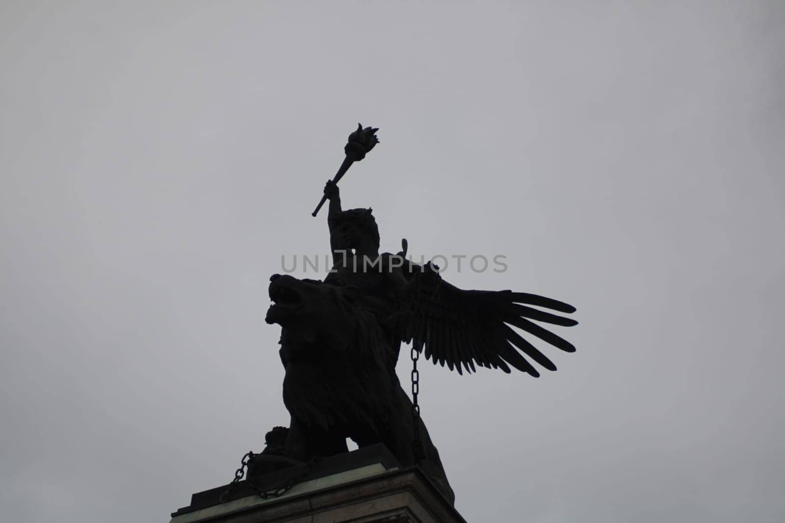 An impressive sculpture depicts an angel with a raised torch, mounted on a lion, silhouetted against a gloomy sky in a serene urban setting.