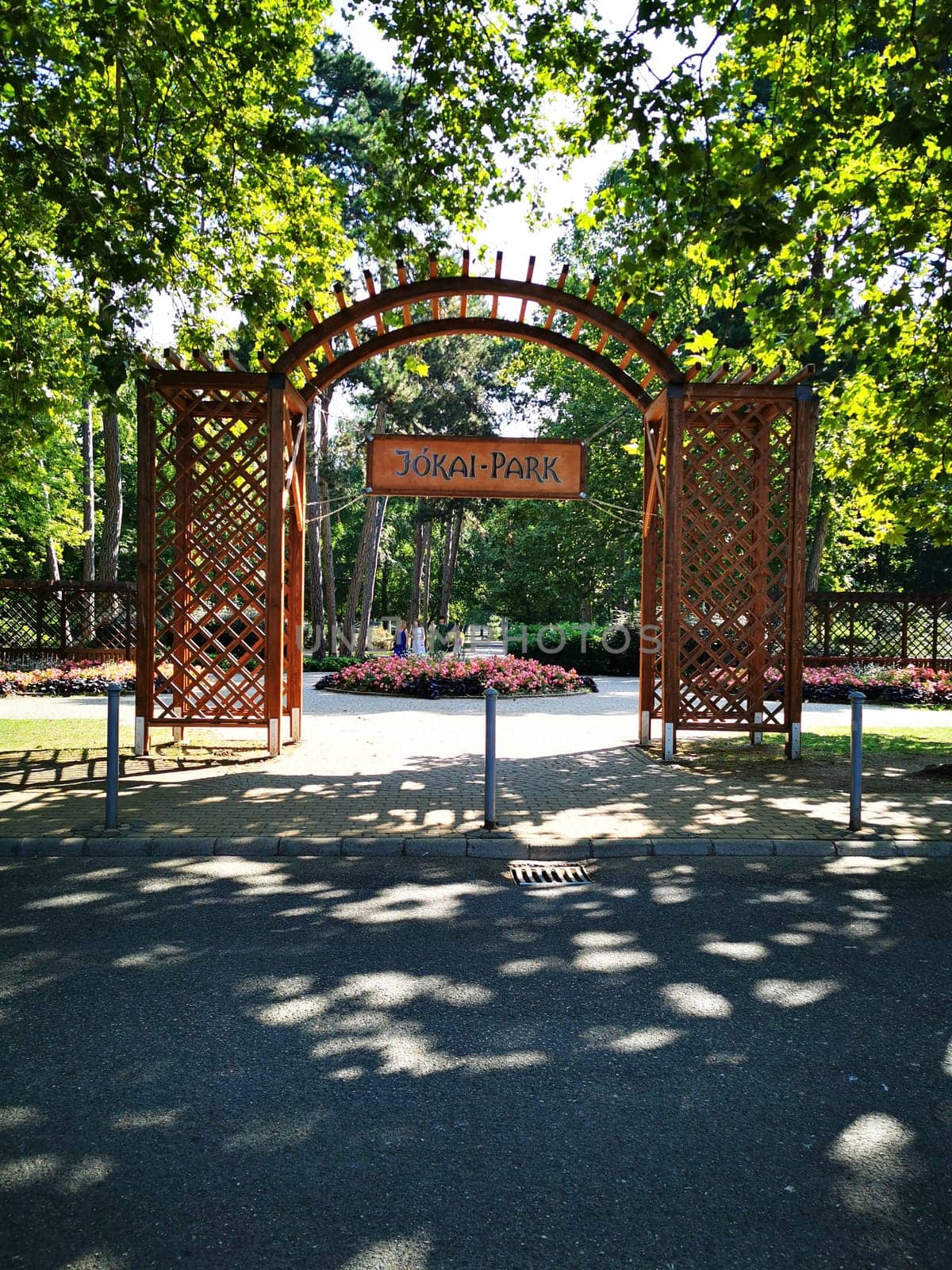 The beautiful entrance to Jókai Park features a decorative archway, leading visitors into a serene space filled with trees and colorful flower beds on a sunny day.