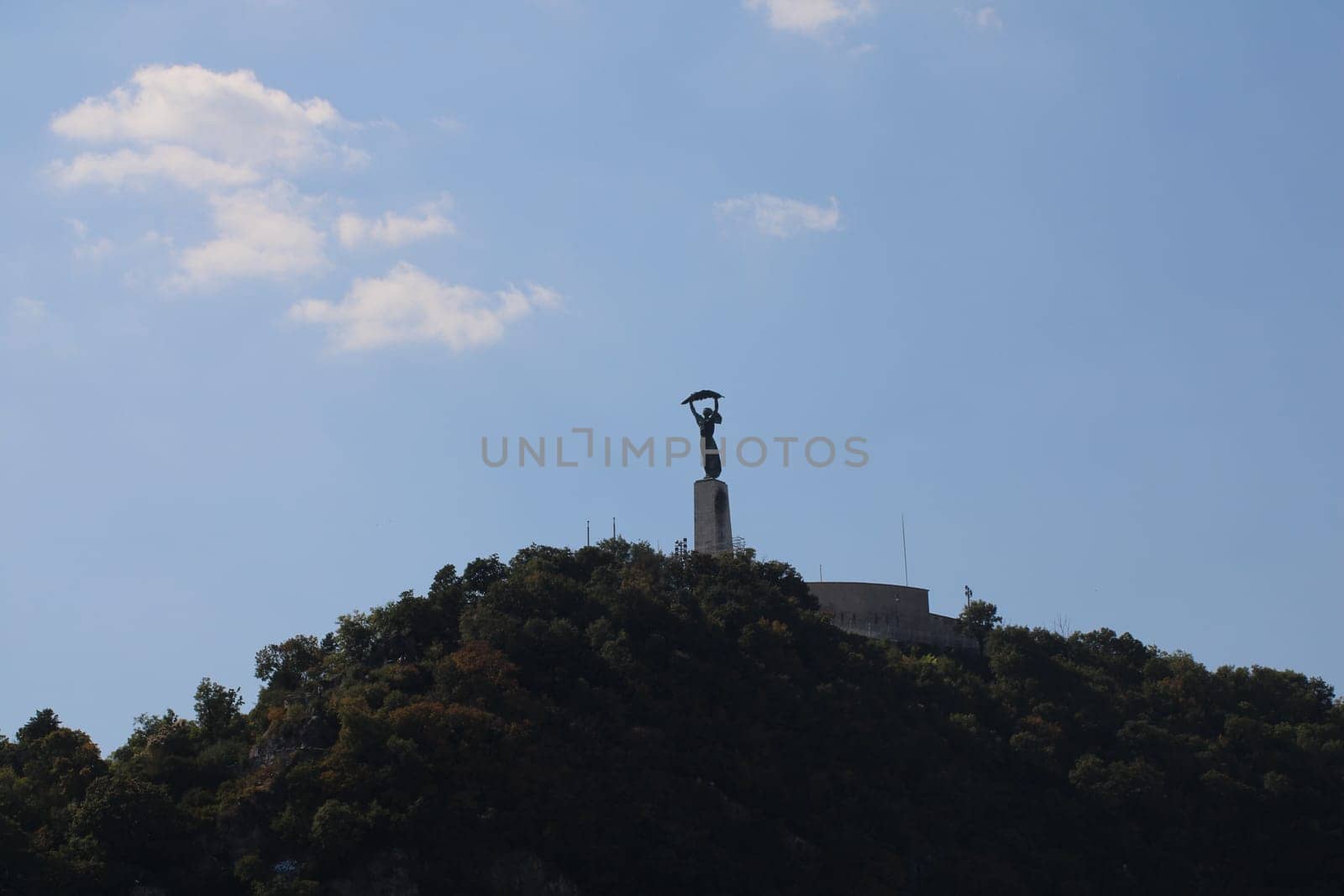 A striking monument of a woman holding an umbrella stands proudly atop a green hill, surrounded by trees, with blue skies and fluffy clouds in the background, indicating a serene daytime atmosphere.