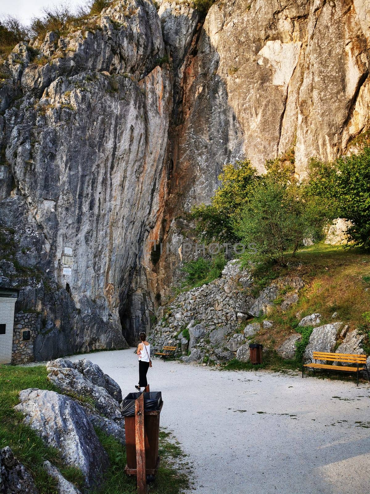 In a tranquil outdoor setting, a person stands at the base of a towering rocky cliff, with lush greenery nearby, enjoying a serene afternoon moment with natural beauty.