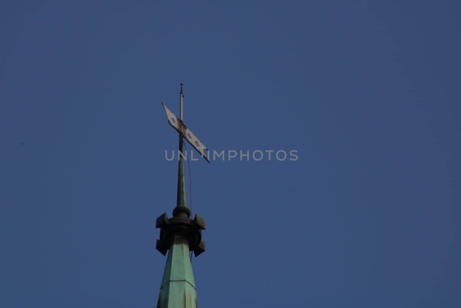 The towering spire of a church stands proudly against the bright blue sky, with a decorative weather vane twirling gently in the midday breeze, showcasing intricate design details.