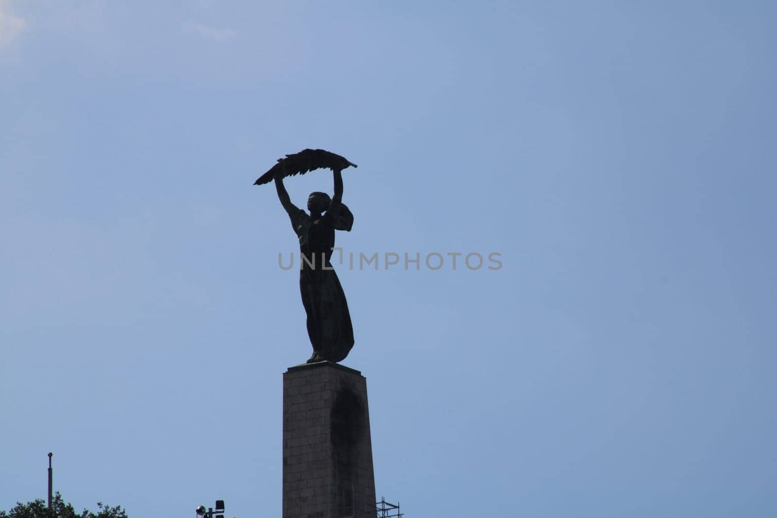 A tall statue depicting a figure holding a bird on a pedestal stands gracefully against a bright blue sky. This serene urban park captures the tranquil essence of the moment.