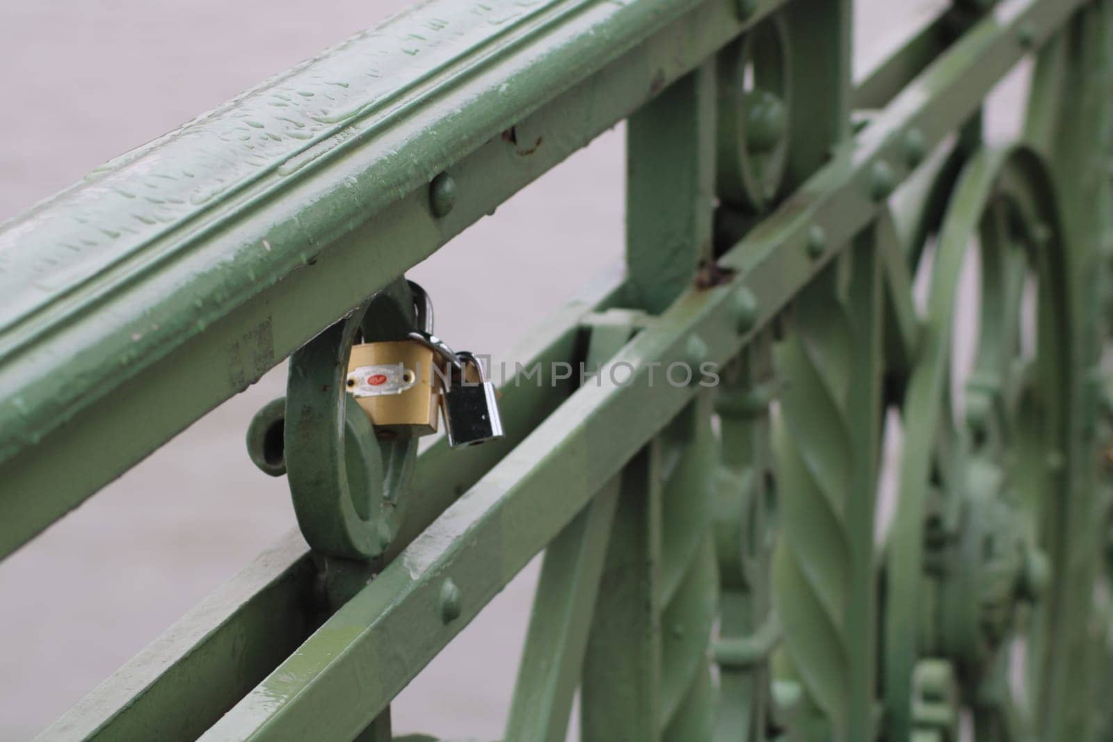 A small padlock attached to a green metal railing on a bridge reflects the tradition of lovers securing their commitments. Light rain adds a serene ambiance to the scene.