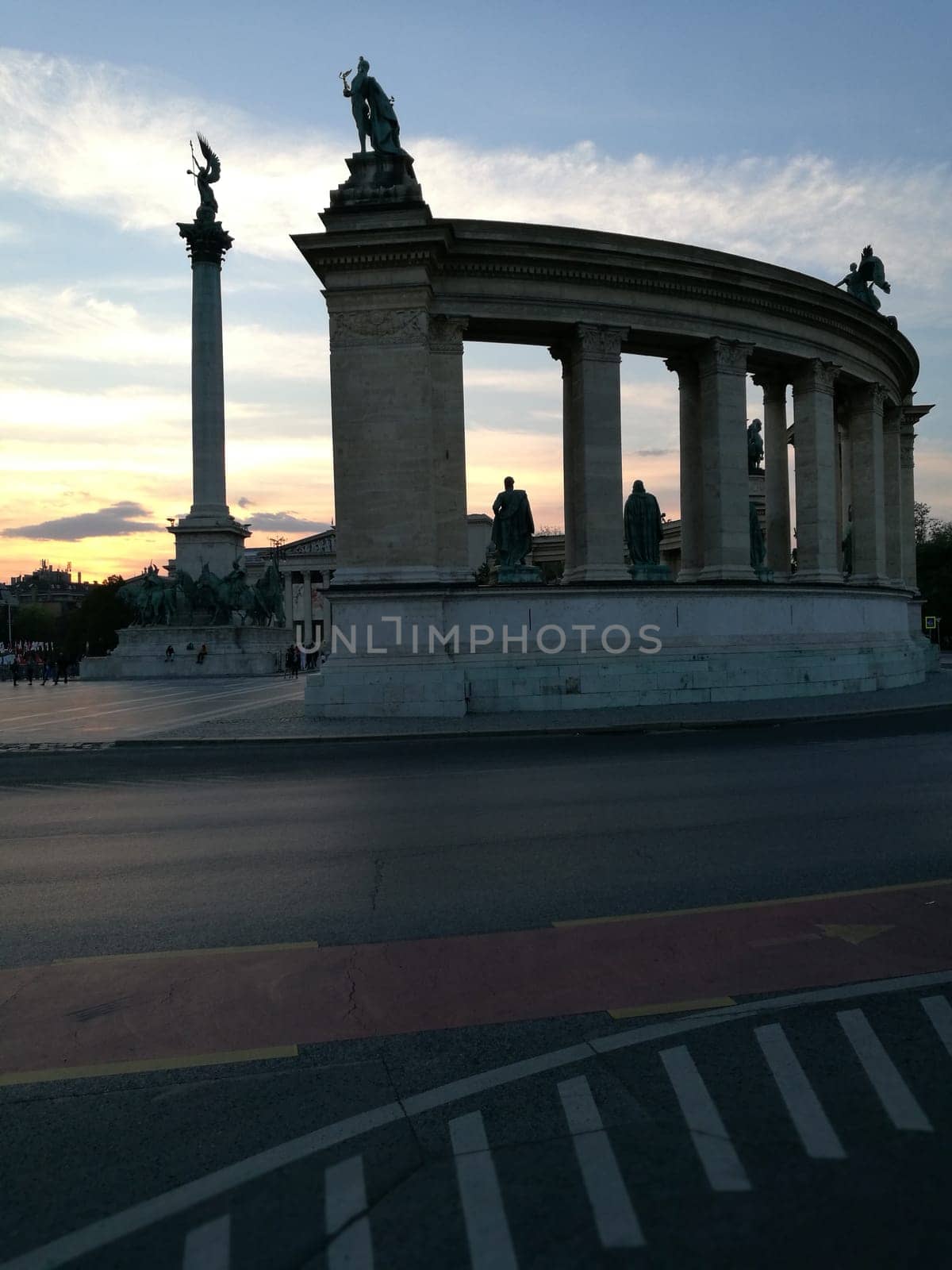 At sunset, a stunning colonnade and monument rise in a lively city square, showcasing intricate statues against a colorful sky, inviting visitors to explore its beauty.