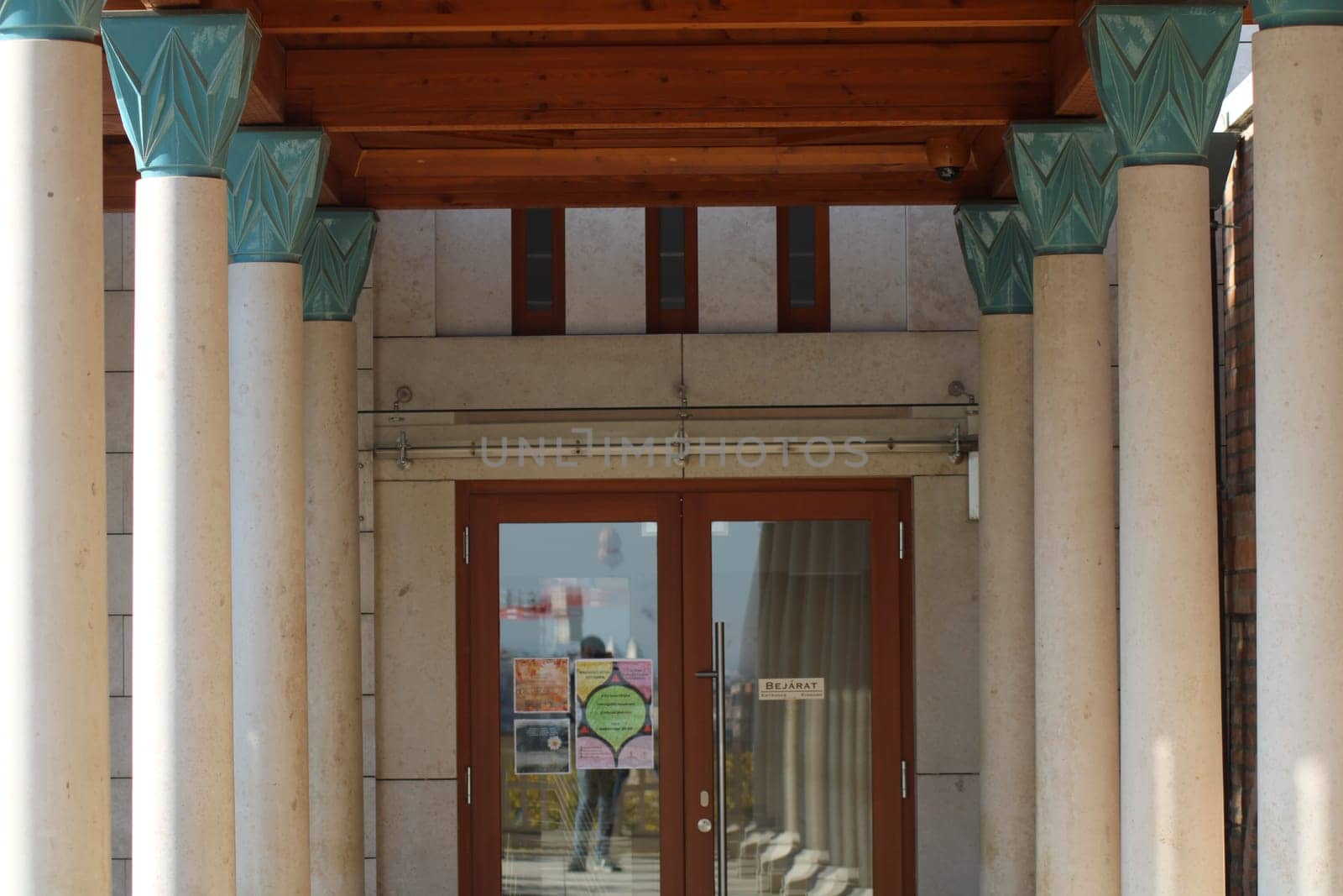The entrance showcases a unique architectural style with tall columns supporting a wooden overhang, inviting visitors into a creative space. The glass doors reflect the surroundings.