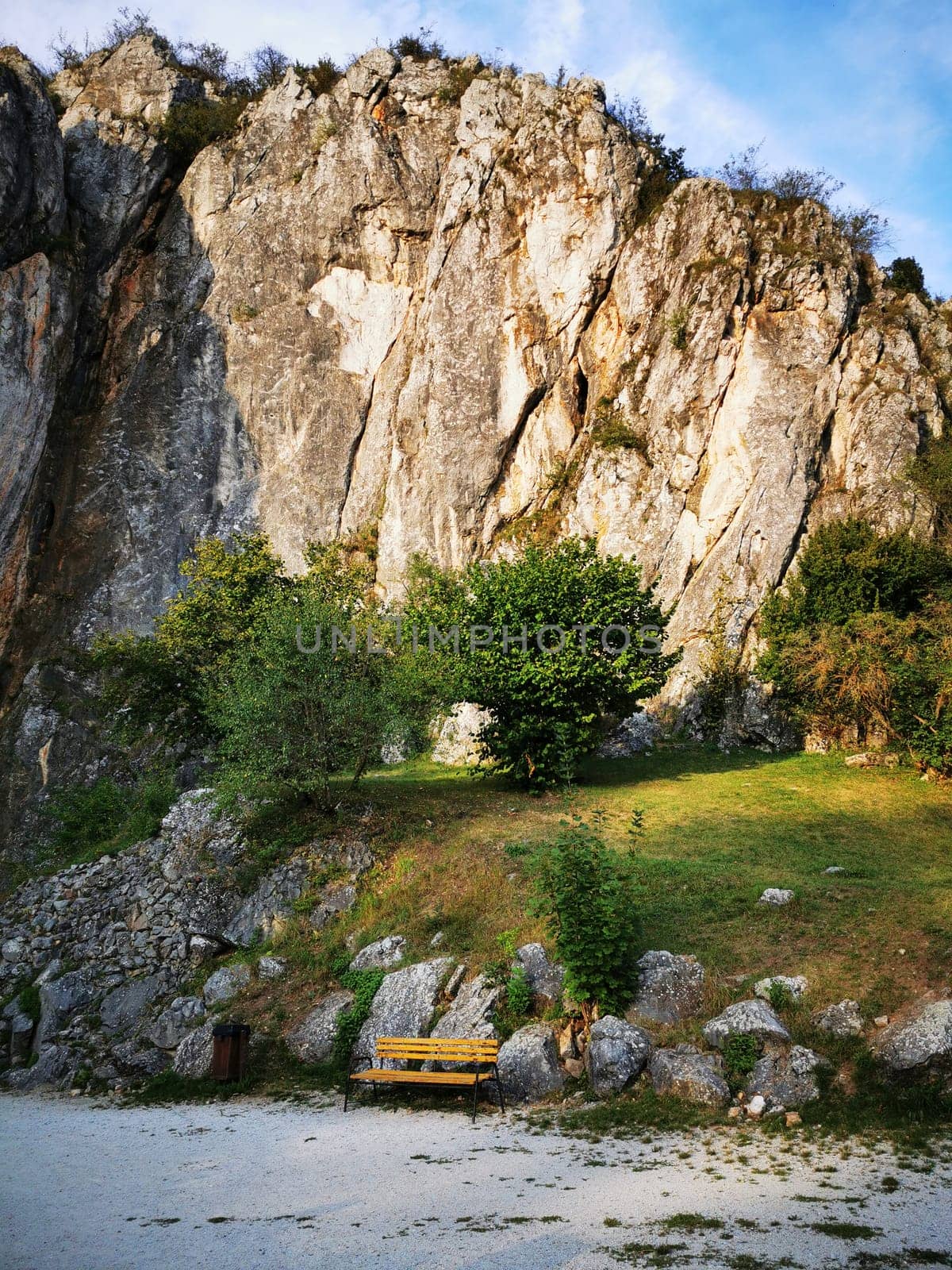 A picturesque view showcases a rugged cliff towering over lush greenery, with a solitary yellow bench resting by the tranquil riverside, perfect for relaxation.