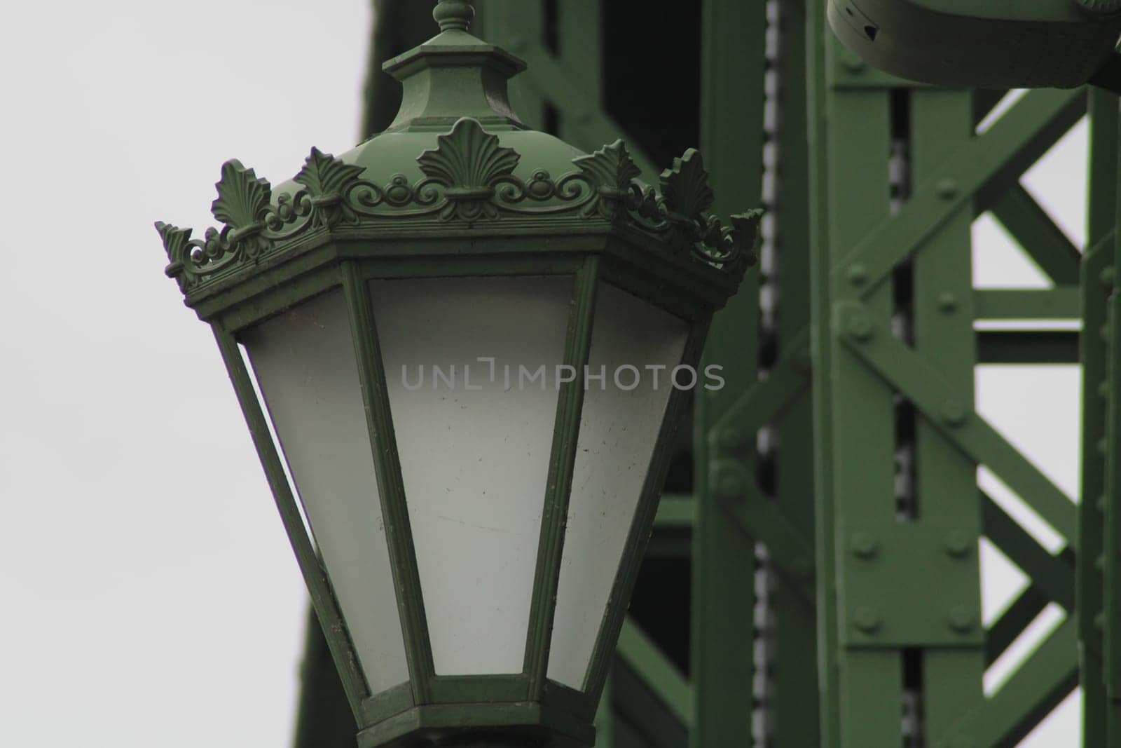 An intricately designed green street lamp stands on a bridge, showcasing decorative elements and ambient lighting, surrounded by industrial structures against a cloudy sky.