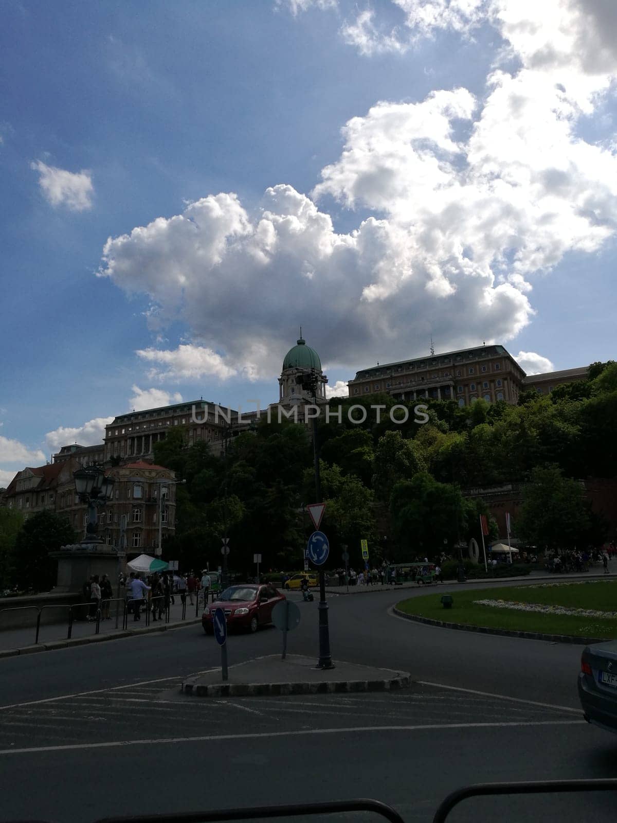 A mix of clouds and blue sky looms over Buda Castle, showcasing its historic architecture and the lush greenery surrounding it on a bright afternoon in Budapest.