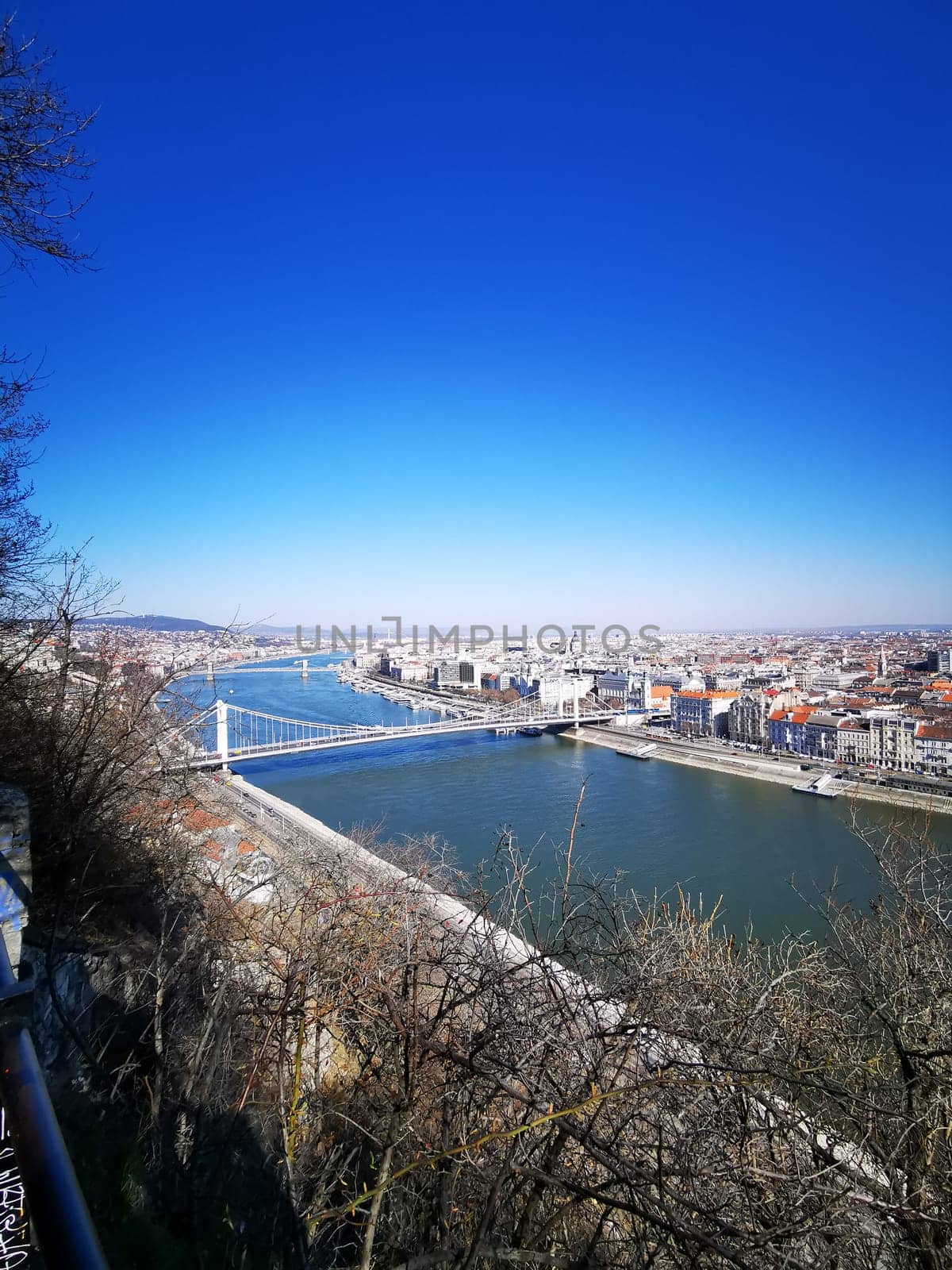 This breathtaking view captures the Danube River flowing through Budapest, revealing iconic bridges and vibrant buildings against a cloudless blue sky.