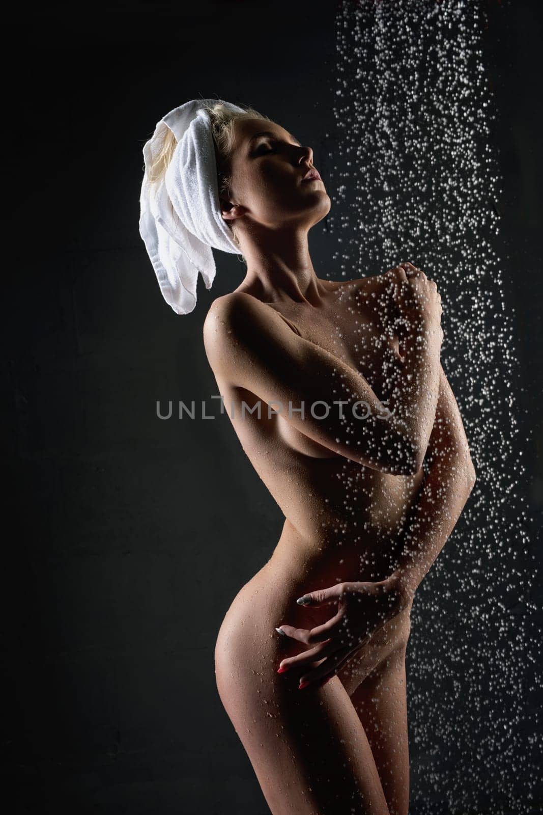 Slim naked blonde with a towel on her head having shower in the dark room cropped view