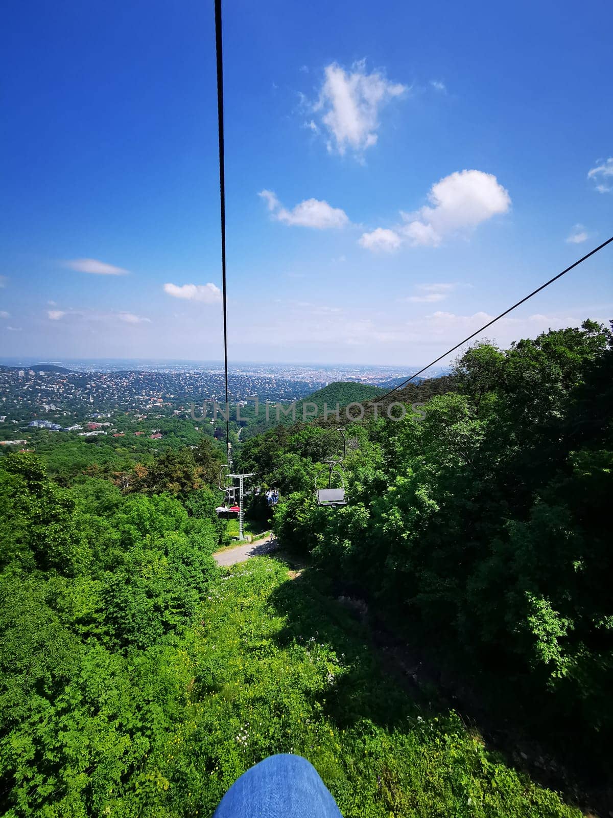 A cable car glides smoothly up the hillside, offering a breathtaking view of the city in the distance beneath a bright blue sky filled with fluffy clouds.