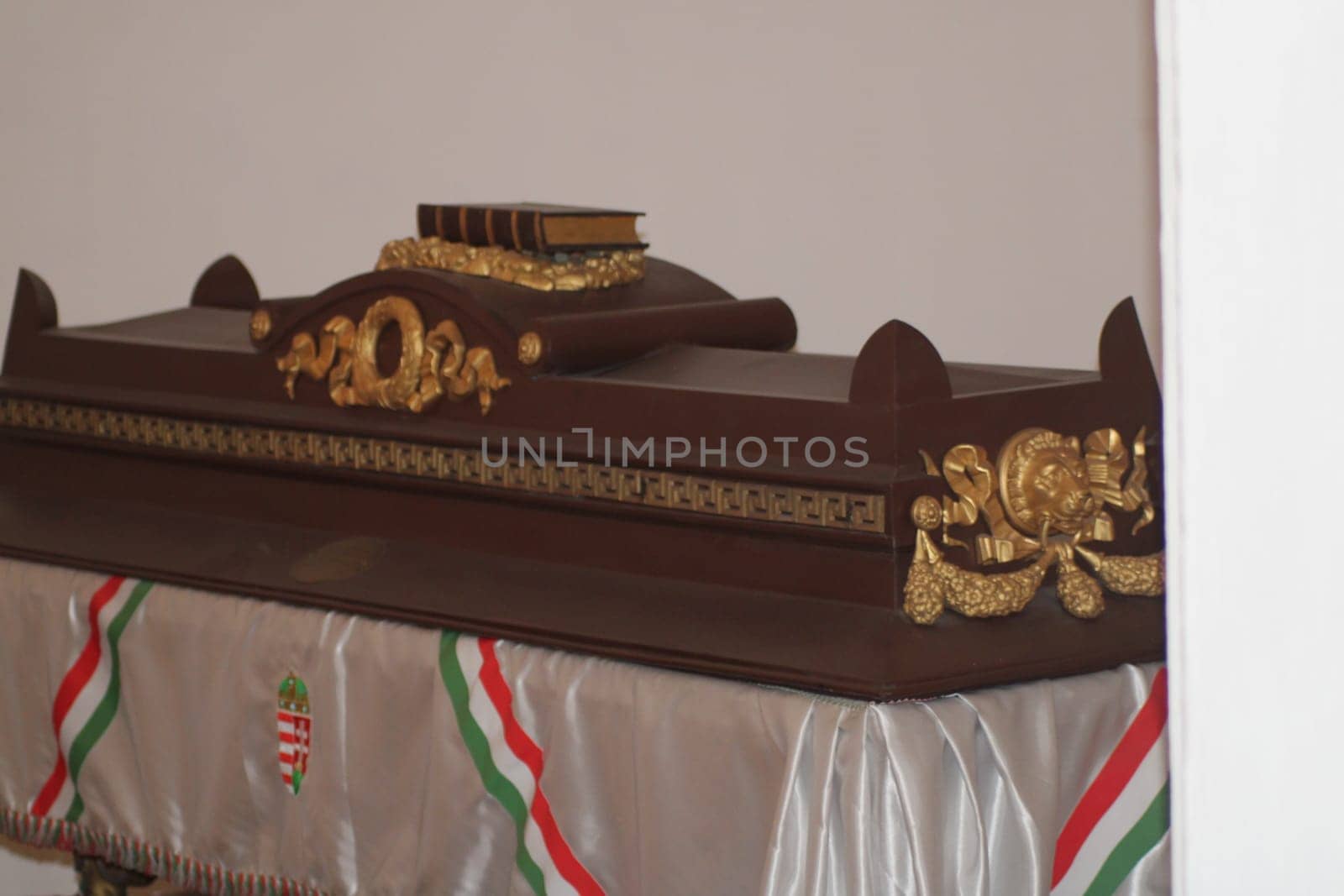 A beautifully crafted wooden coffin with gold accents rests in a quiet space, adorned with a decorative cloth featuring colorful ribbons, creating a respectful atmosphere.