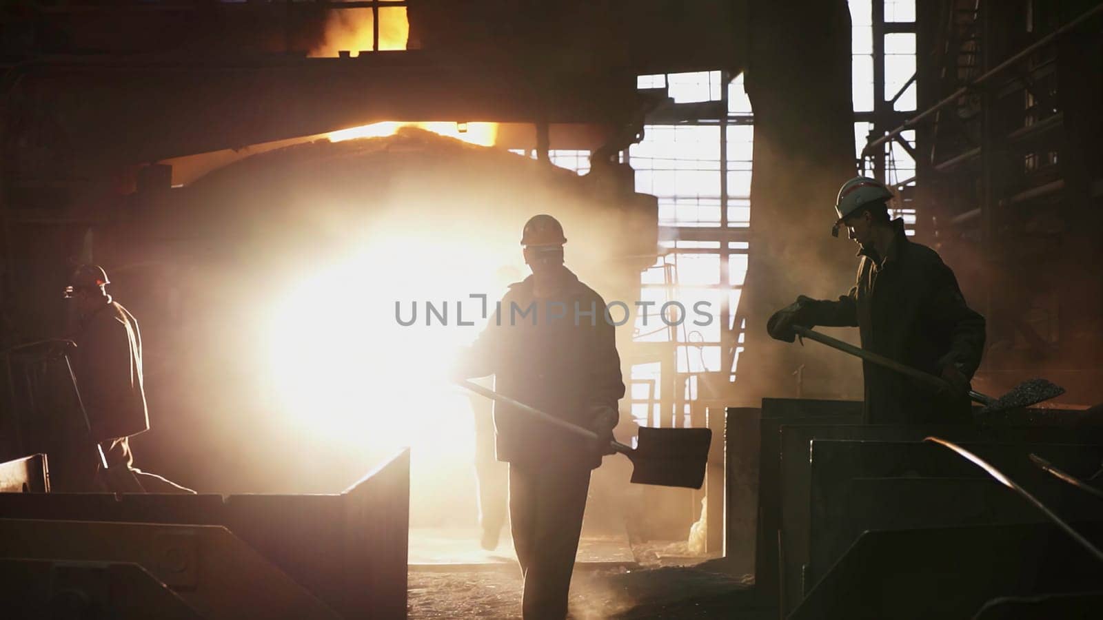 Silhouetted workers diligently laboring in a bustling steel mill, with molten metal glowing intensely in the background. Stock Clip