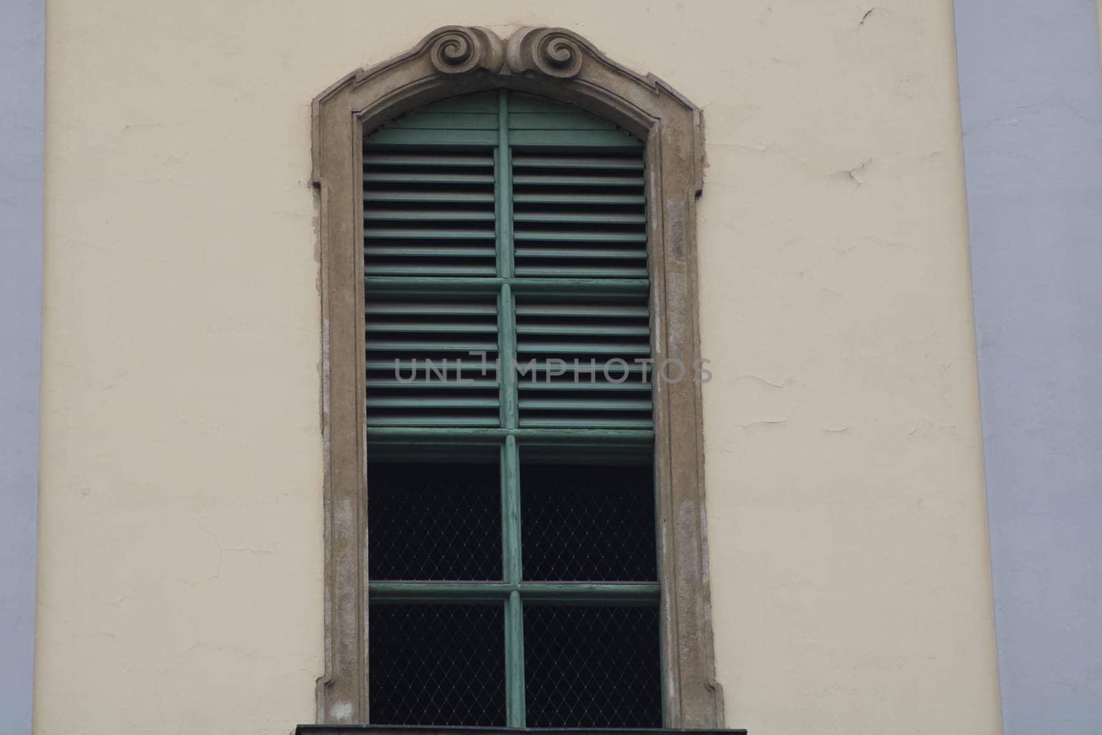 An ornate window featuring green wooden shutters is set within a weathered building wall. The intricate design adds character to the architecture in bright sunlight.