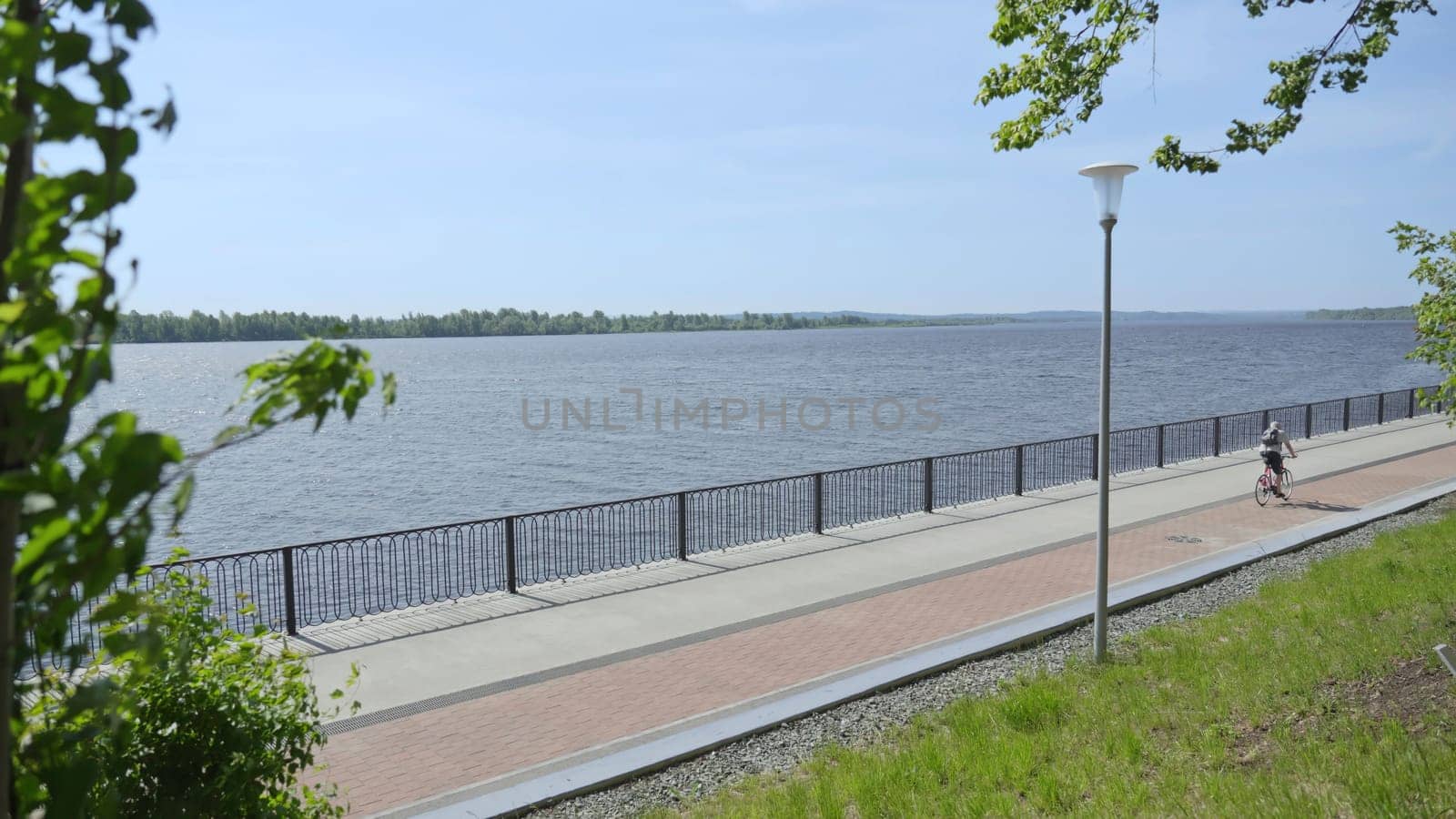 On this wonderful day, a cyclist is riding by the scenic waterfront, surrounded by lush greenery and bright blue skies. Clip