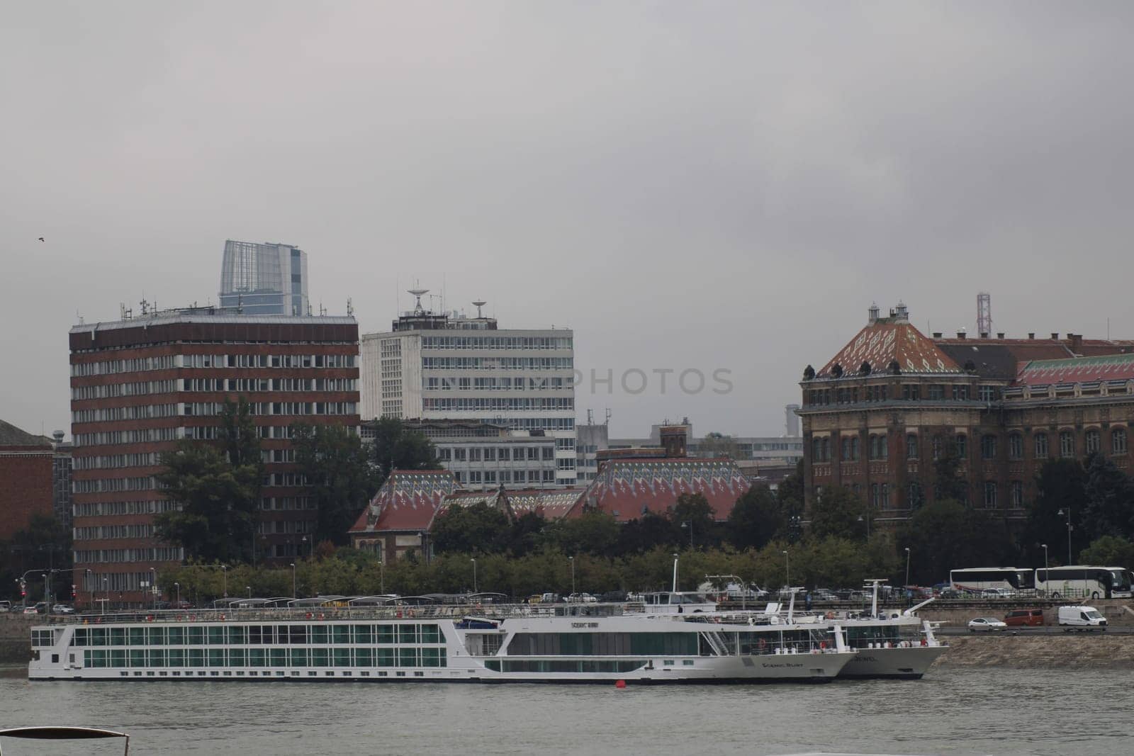 A river cruise ship sails along the Danube River in Budapest, surrounded by a mix of modern and historical buildings under overcast skies.