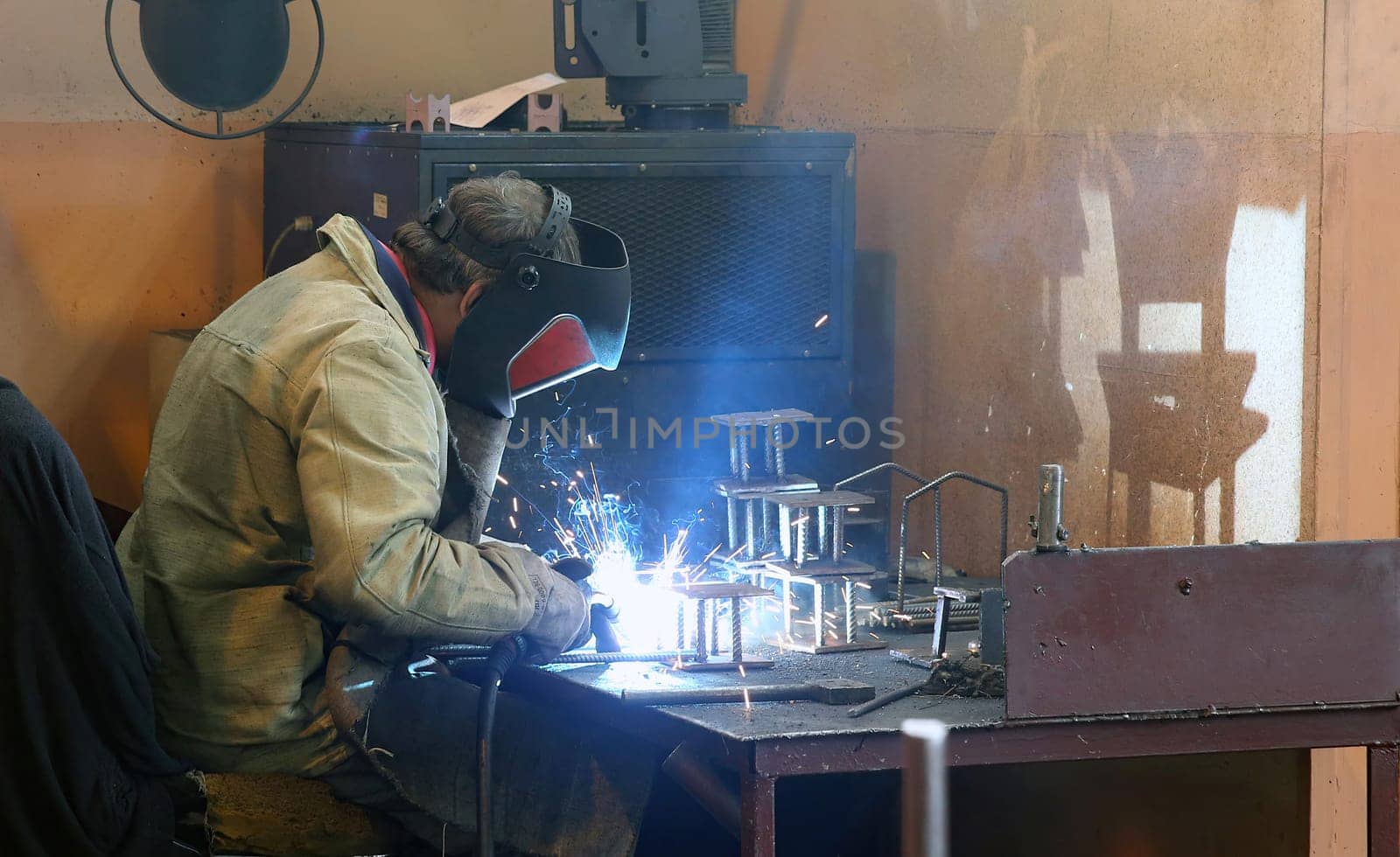 A welder, wearing a protective mask and gloves, focuses intently on welding metal pieces together, creating sparks and a bright light in a workshop setting.