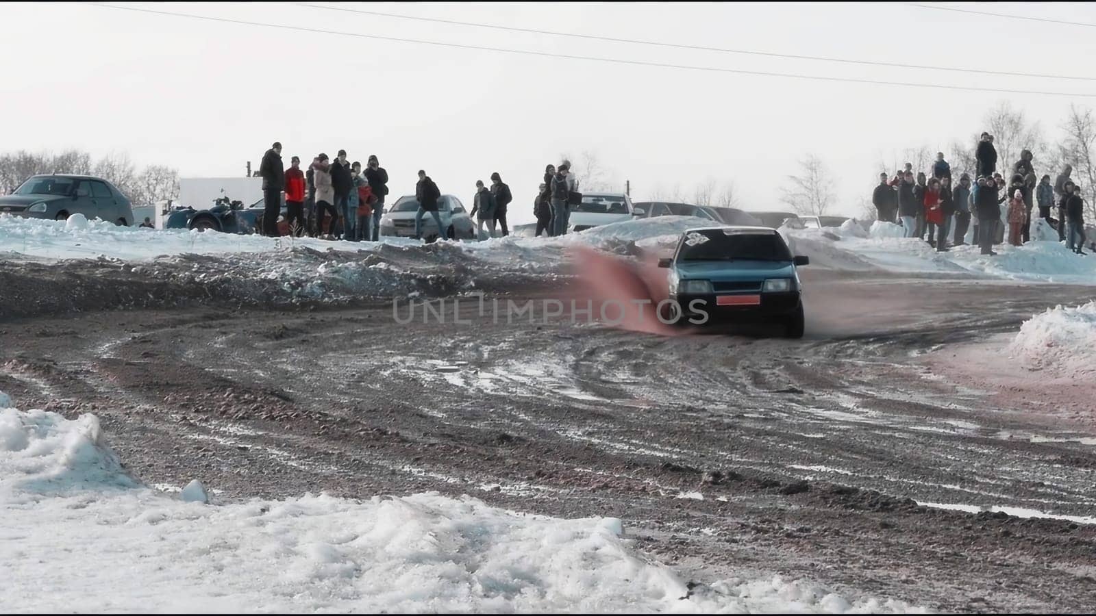 Experience winter driving as a skilled driver navigates through snow, drifting gracefully on a dirt road, impressing a captivated crowd nearby
