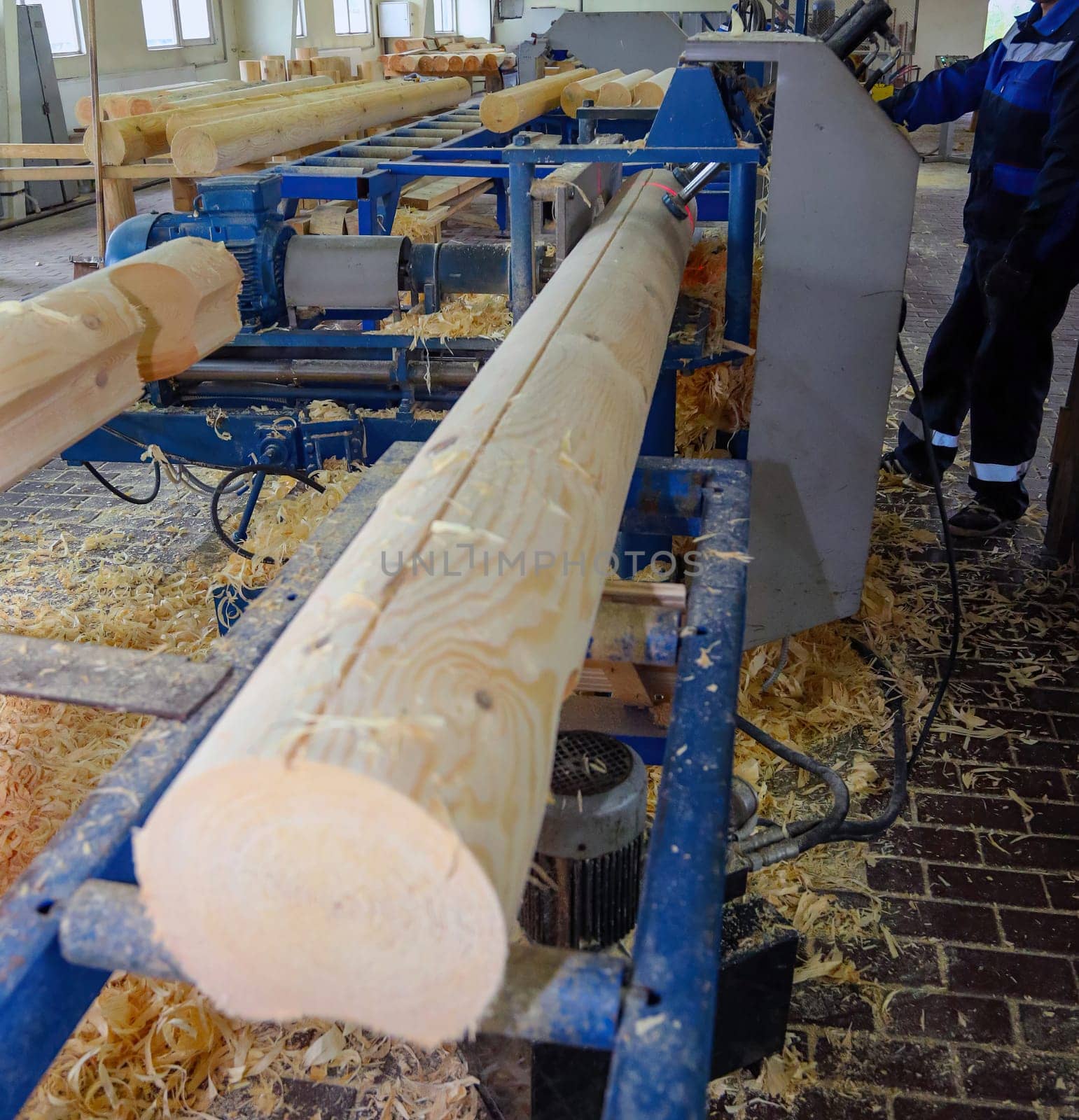 A worker stands at a wood processing machine, shaping logs from rough timber into smooth pieces in a busy workshop.