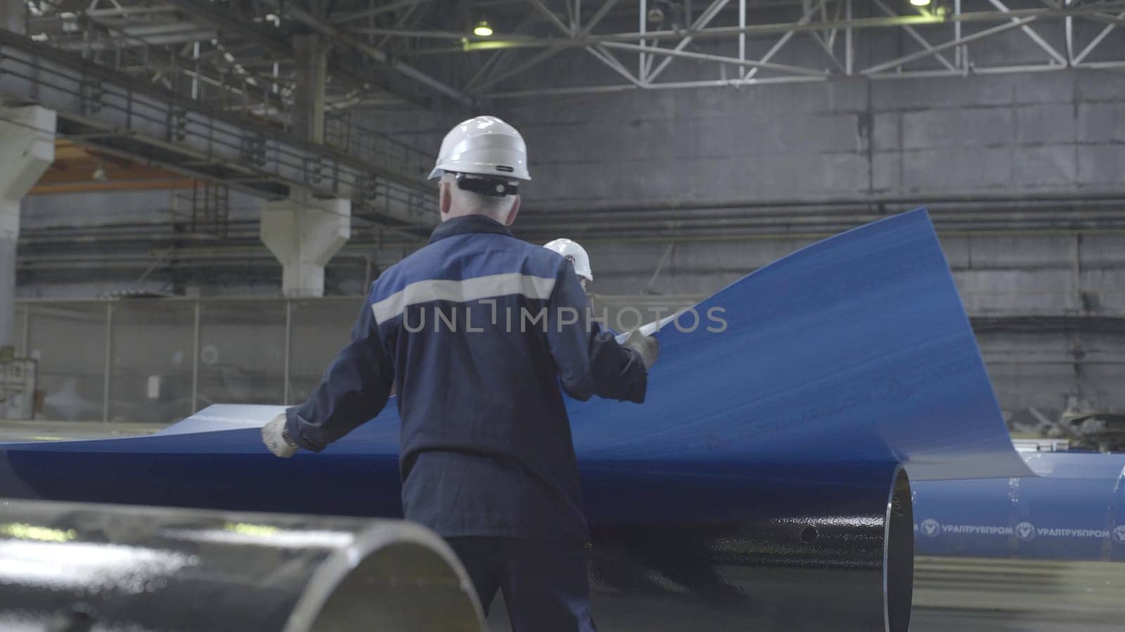 This video shows a large warehouse filled with metal pipes, featuring a skilled worker in a hard hat handling various pieces, including a blue pipe