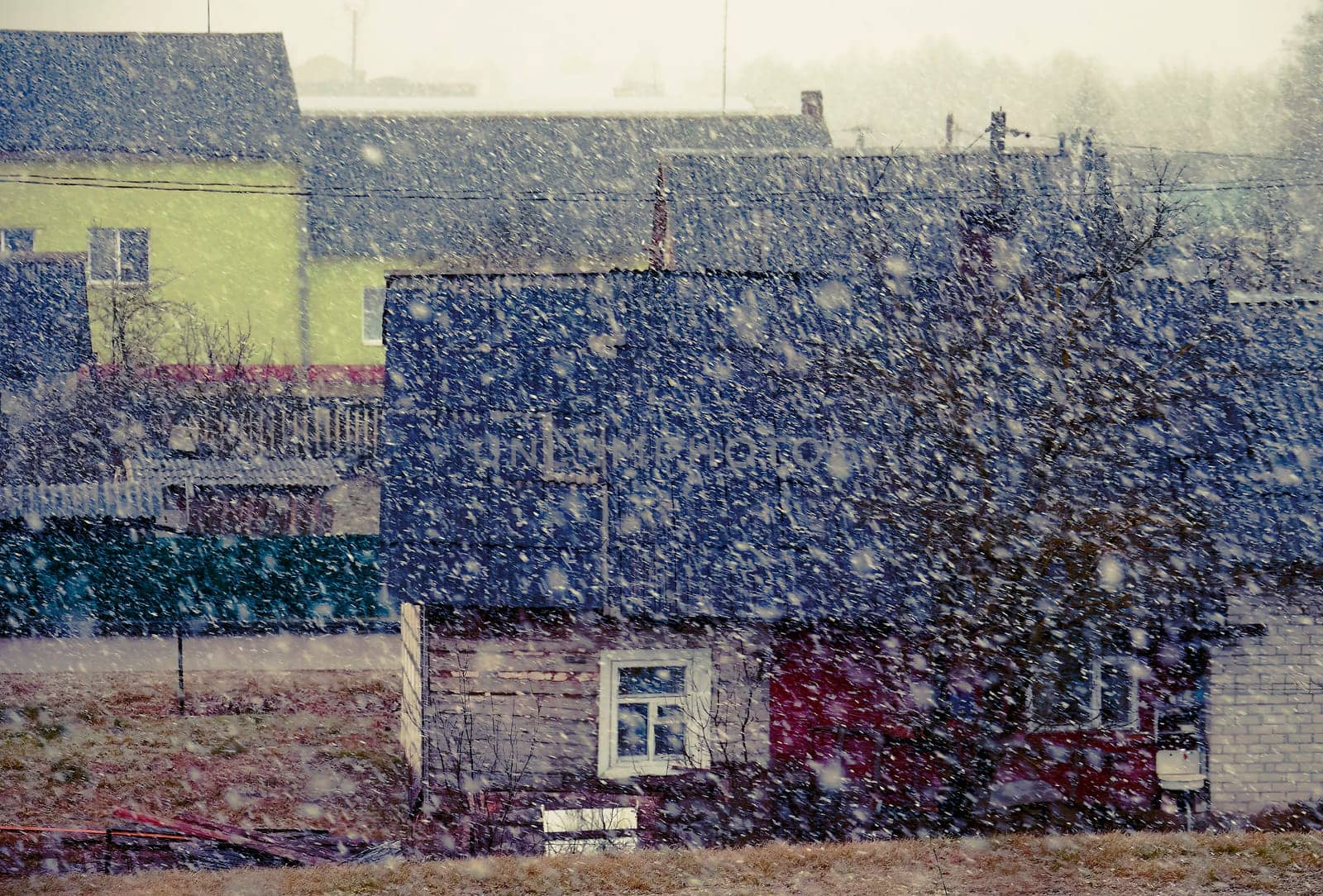 The image shows a small village on a gloomy winter day. The snow is falling heavily, and the houses are covered in snow. The image has a cold and desolate feel to it.