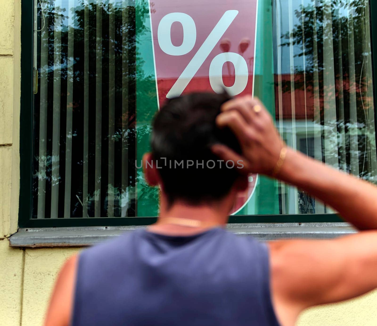 A man is standing outside a store, scratching his head as he observes a large percentage sign displayed in the window. His posture suggests he might be puzzled or considering the implications of the advertised discount, sale, or promotion.