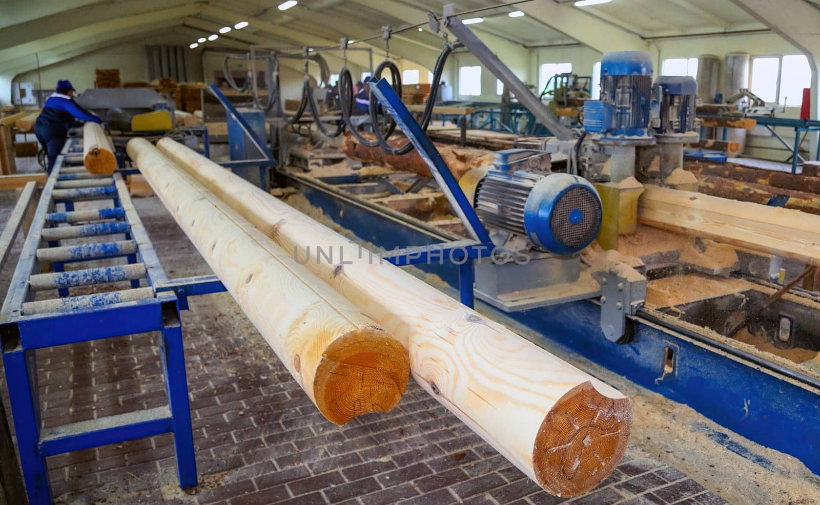 Workers are actively cutting large logs into planks at a lumber mill, highlighting machining equipment and wood shavings on the factory floor.