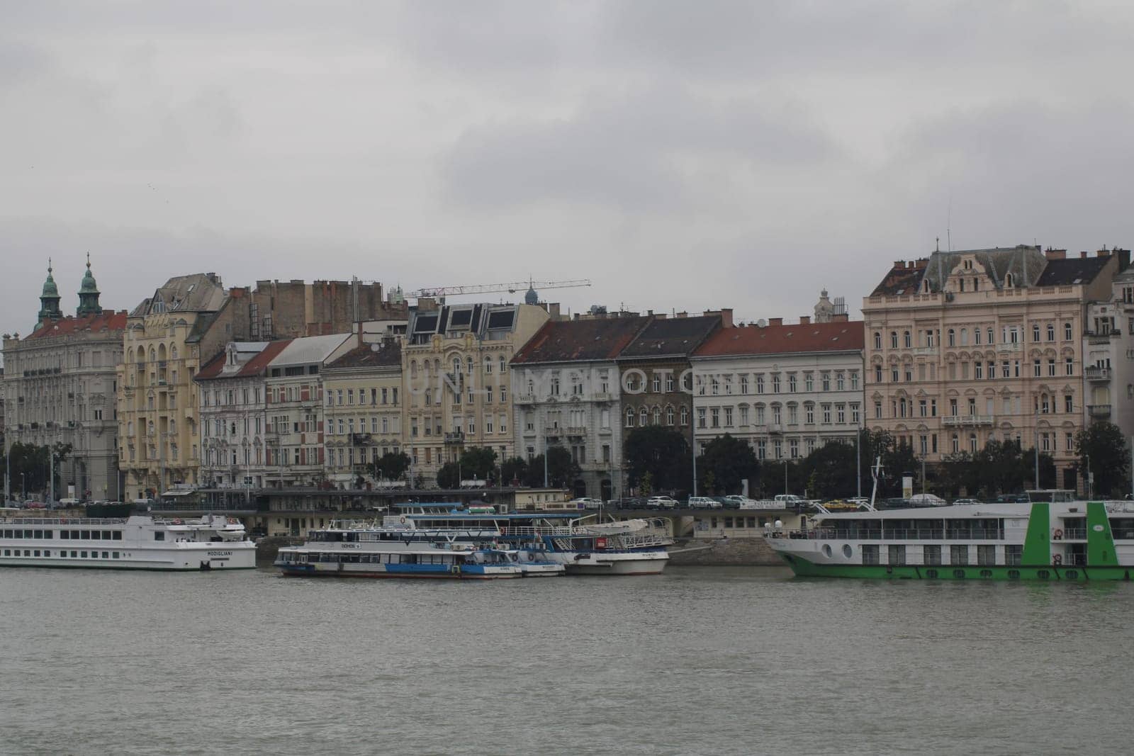 Several boats are moored along the river in Budapest, surrounded by beautiful historical architecture under an overcast sky.