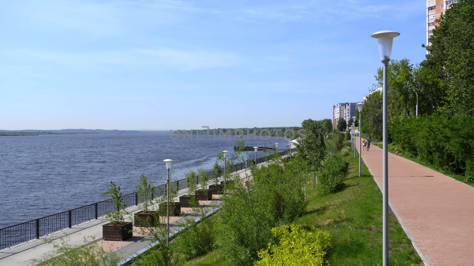 A peaceful and serene riverside walkway is beautifully surrounded by lush greenery beneath a clear blue sky. Clip