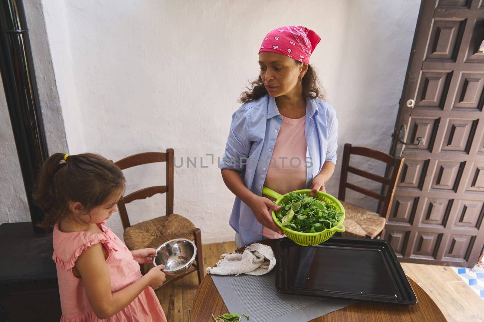 A mother and daughter spend quality time together in the kitchen, preparing a fresh green salad. The mother holds a colander filled with leafy greens while the daughter helps with a bowl.