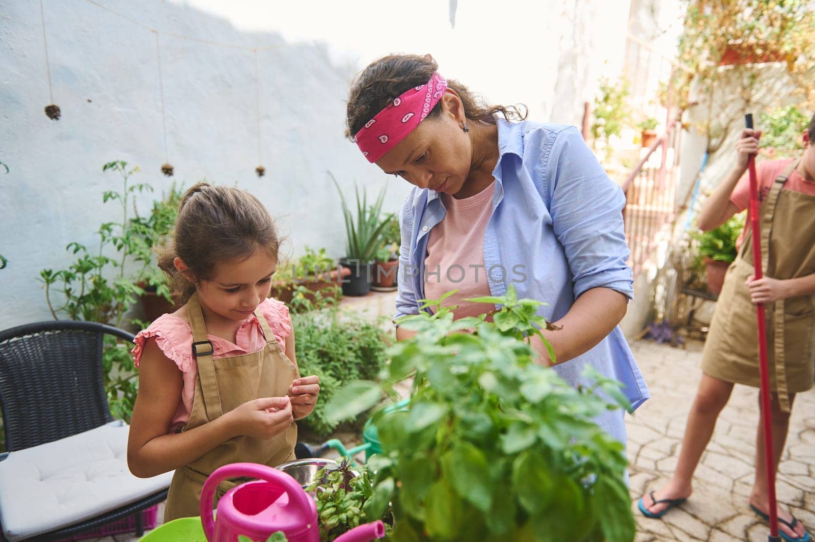 A mother and daughter enjoy gardening together on a sunny patio, surrounded by lush plants. The girl focuses intently while the mother guides her, creating a warm family bonding moment.