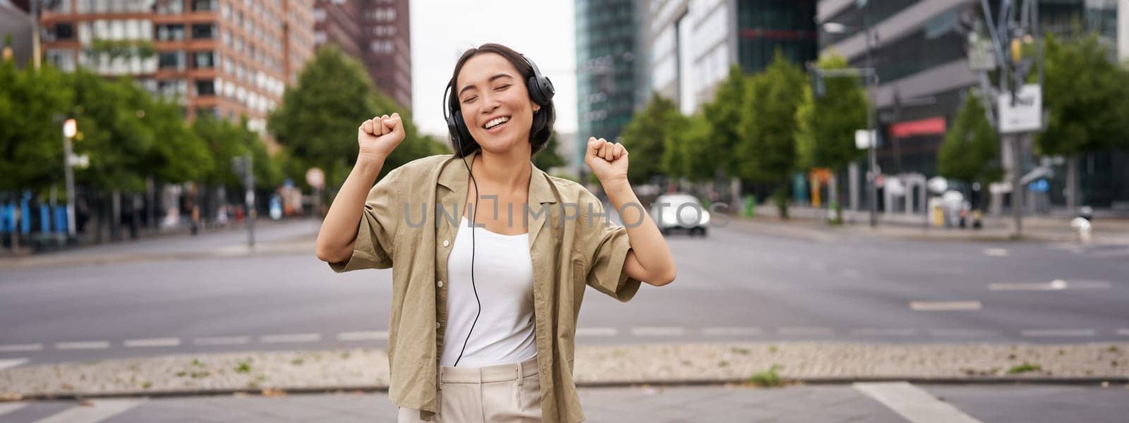 Dancing girl feeling happy in city. Asian woman dancing and listening music in headphones, posing on street by Benzoix